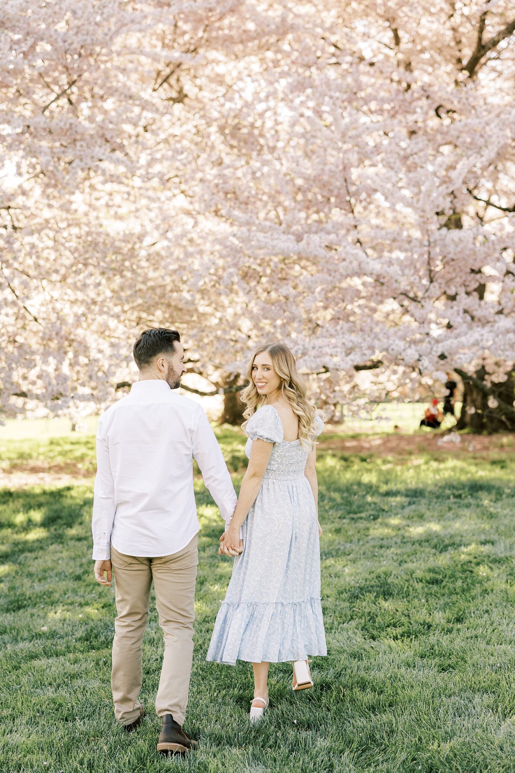 engaged couple holds hands walking through cherry blossom trees
