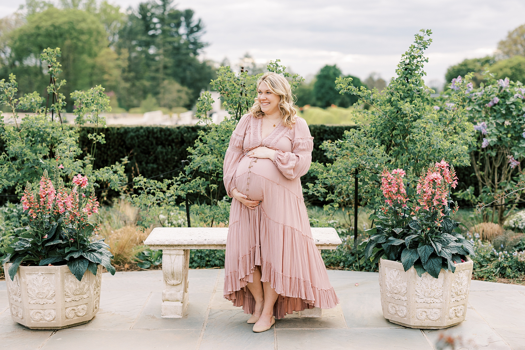 mom stands by bench between pink flowers in pots holding her baby bump during maternity photos in Philadelphia