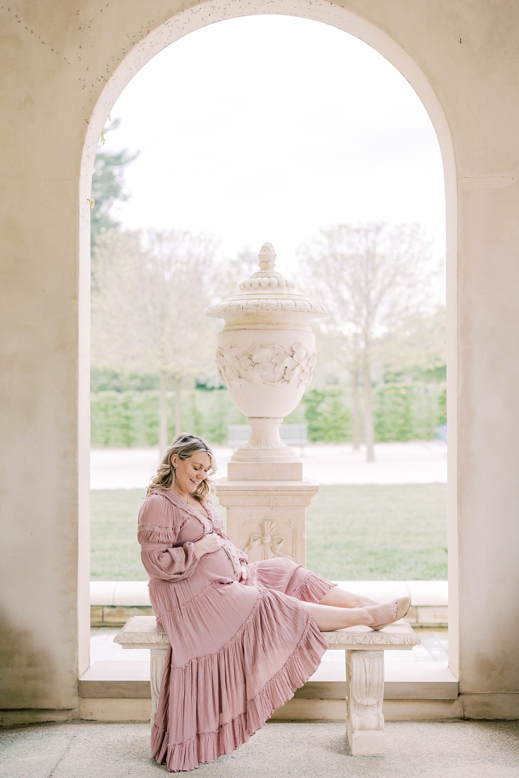mom in pink dress sits on bench in Longwood Gardens holding baby bump