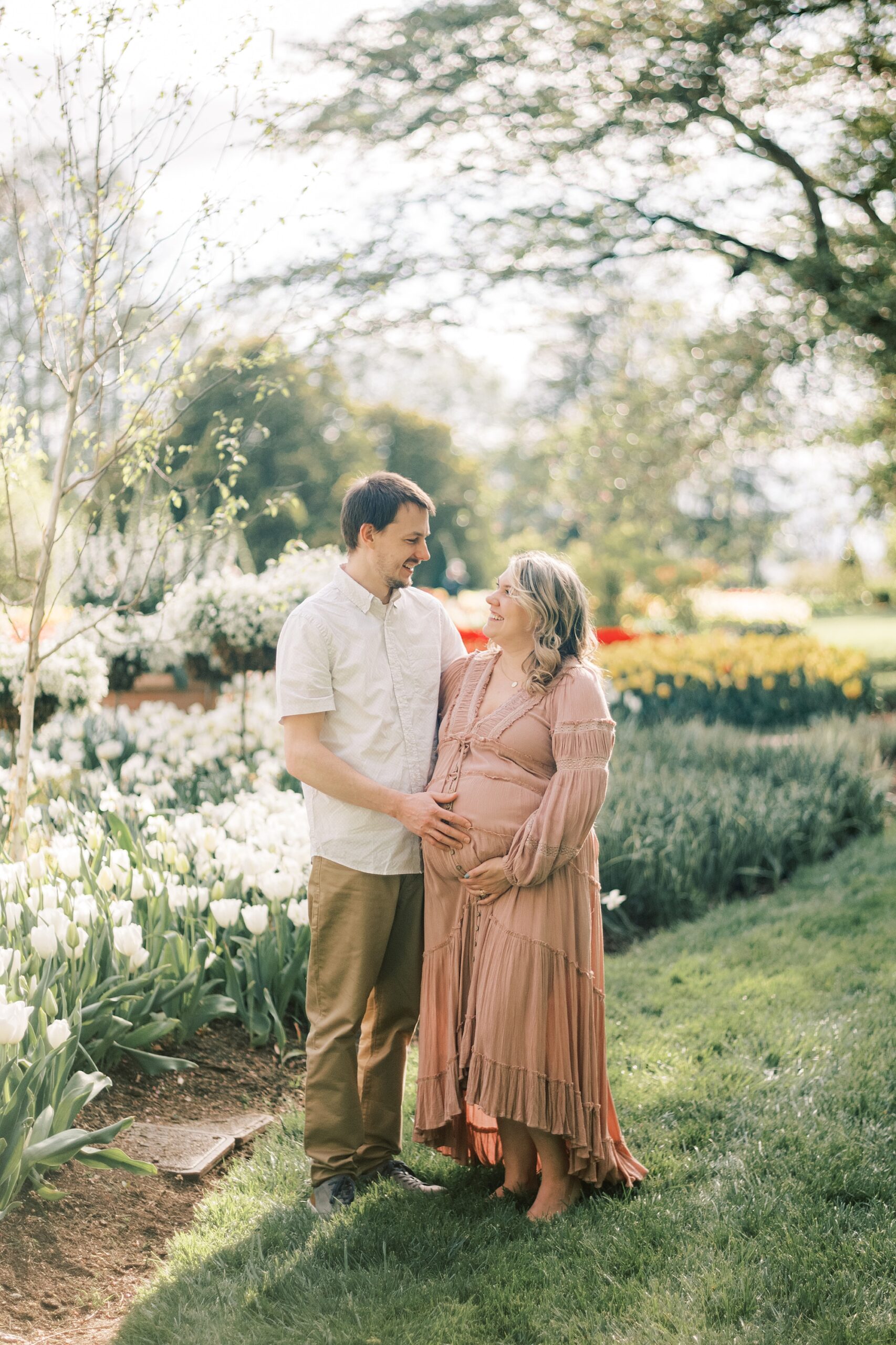 expecting parents hold mom's baby bump by flower display during maternity photos in Philadelphia