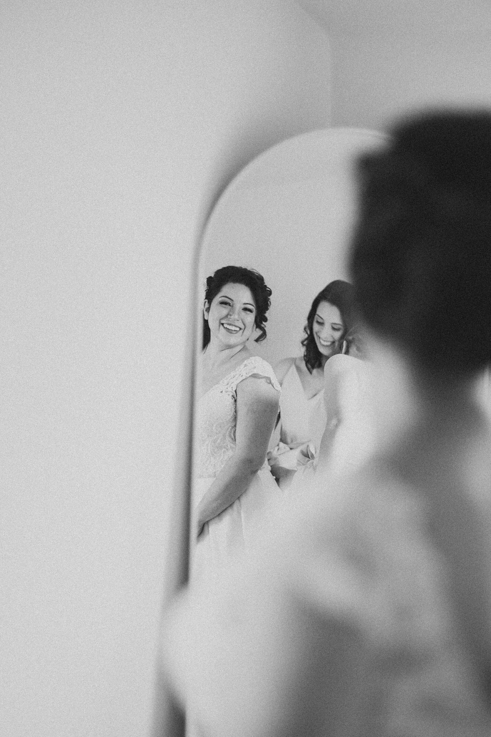 woman helps bride into wedding dress looking in mirrored reflection 