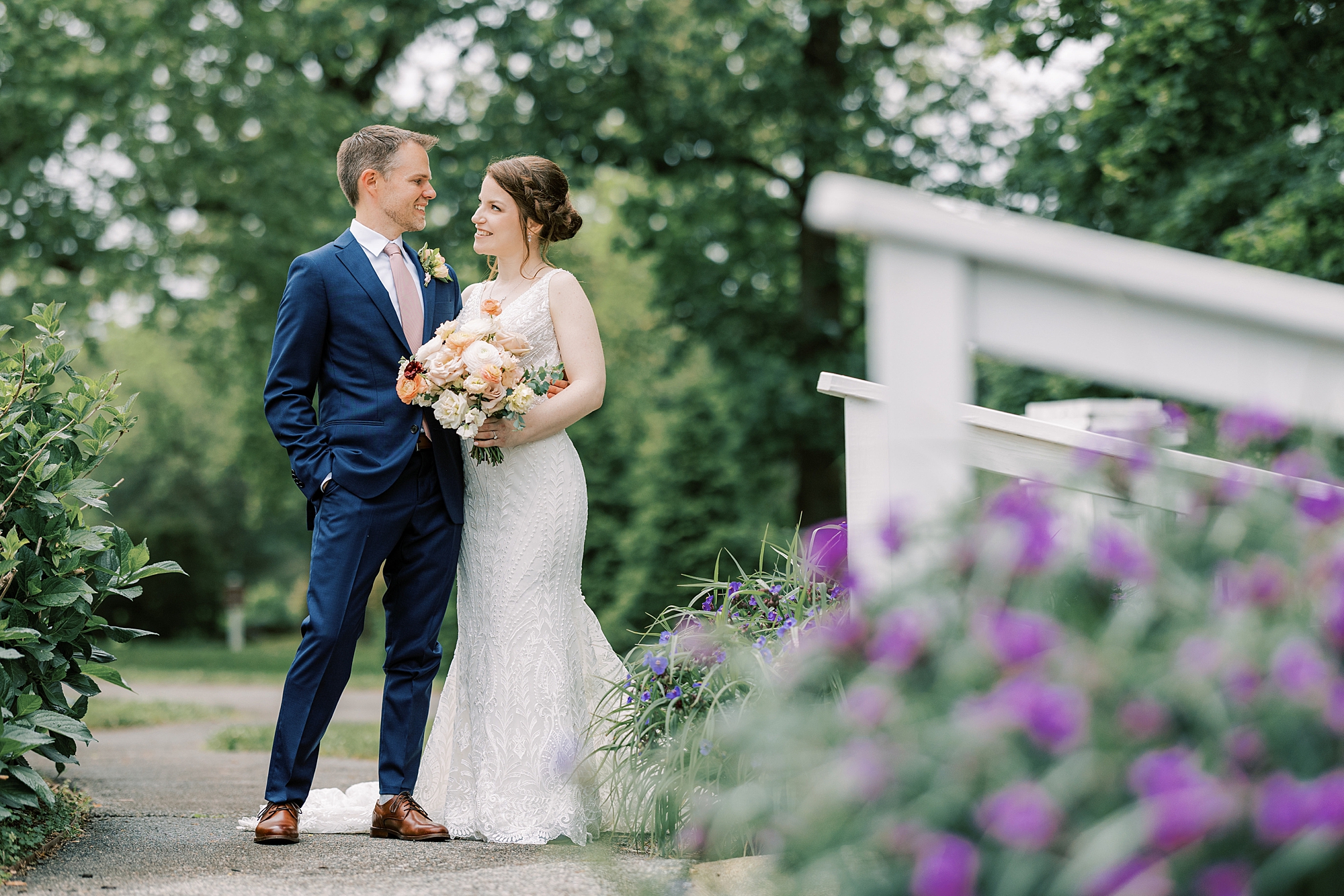 bride and groom smile on bridge with purple flowers in foreground 