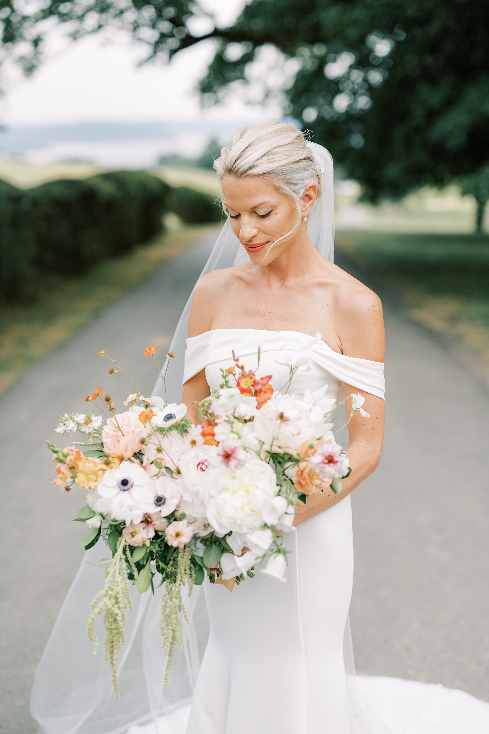 blonde bride in off-the-shoulder gown looks down at pastel bouquet