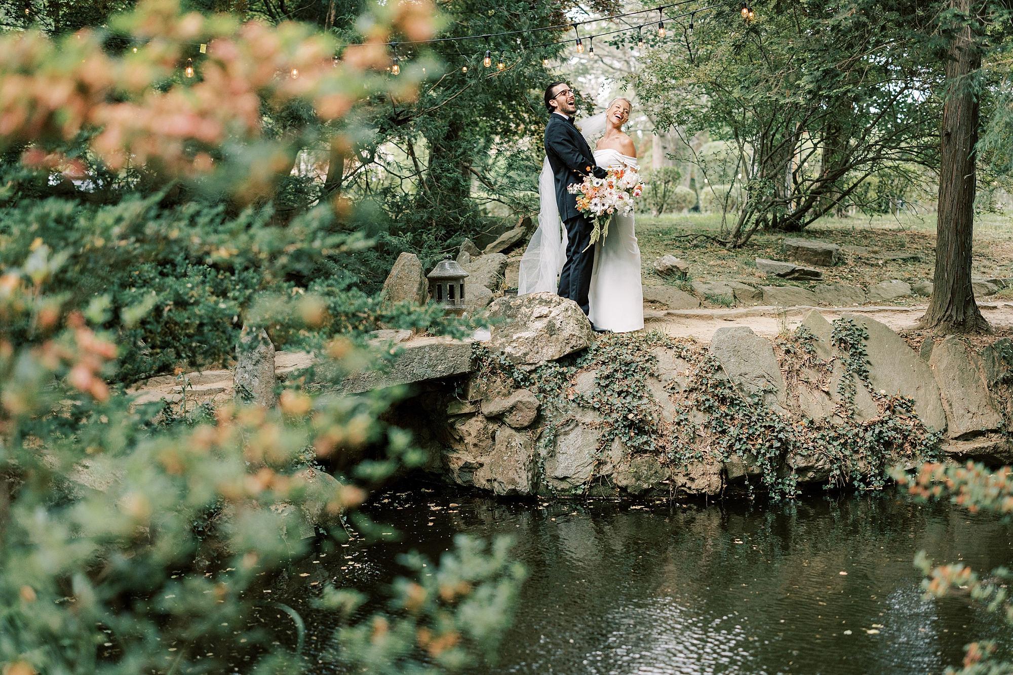 bride and groom pose on stone wall overlooking pond in Japanese Gardens of Lauxmont Farms