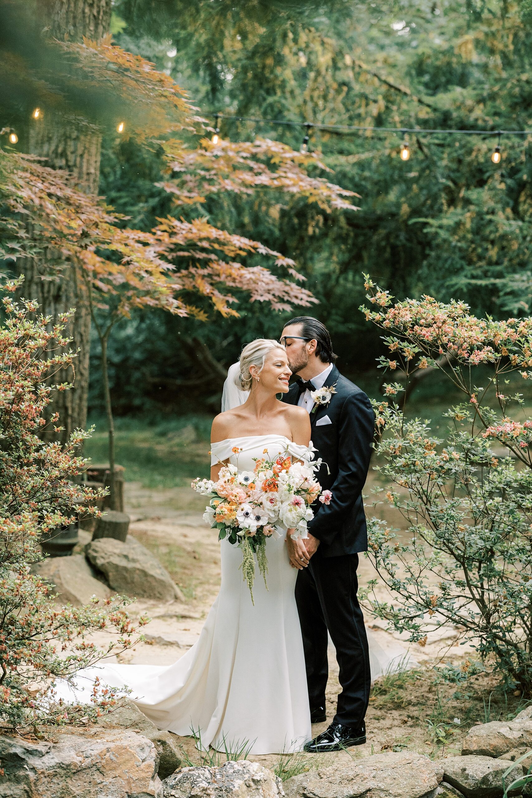groom kisses bride's cheek on stone wall in Japanese Gardens of Lauxmont Farms