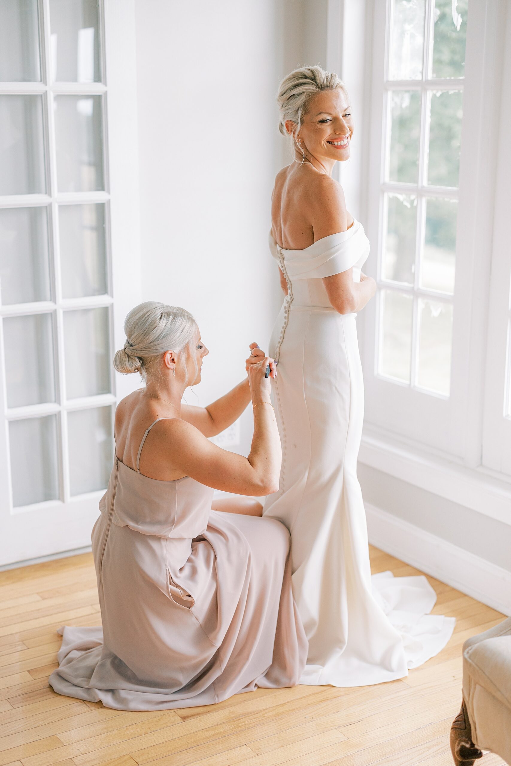 bridesmaid in pink gown helps bride with buttons on wedding gown