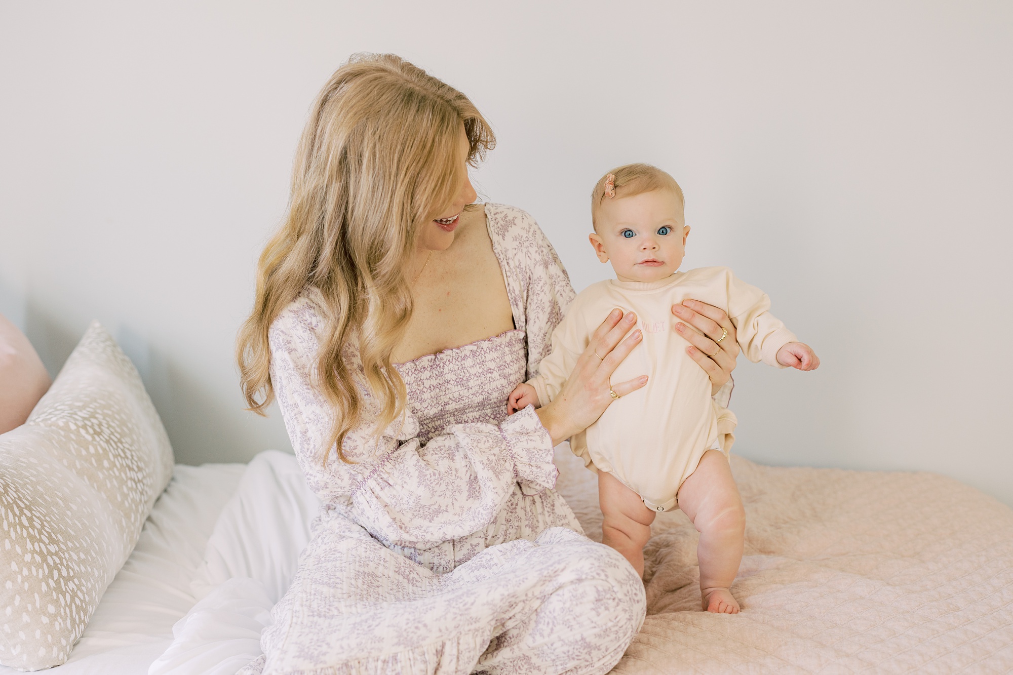 mom in pink and white gown holds up toddler on bed during lifestyle photos at home
