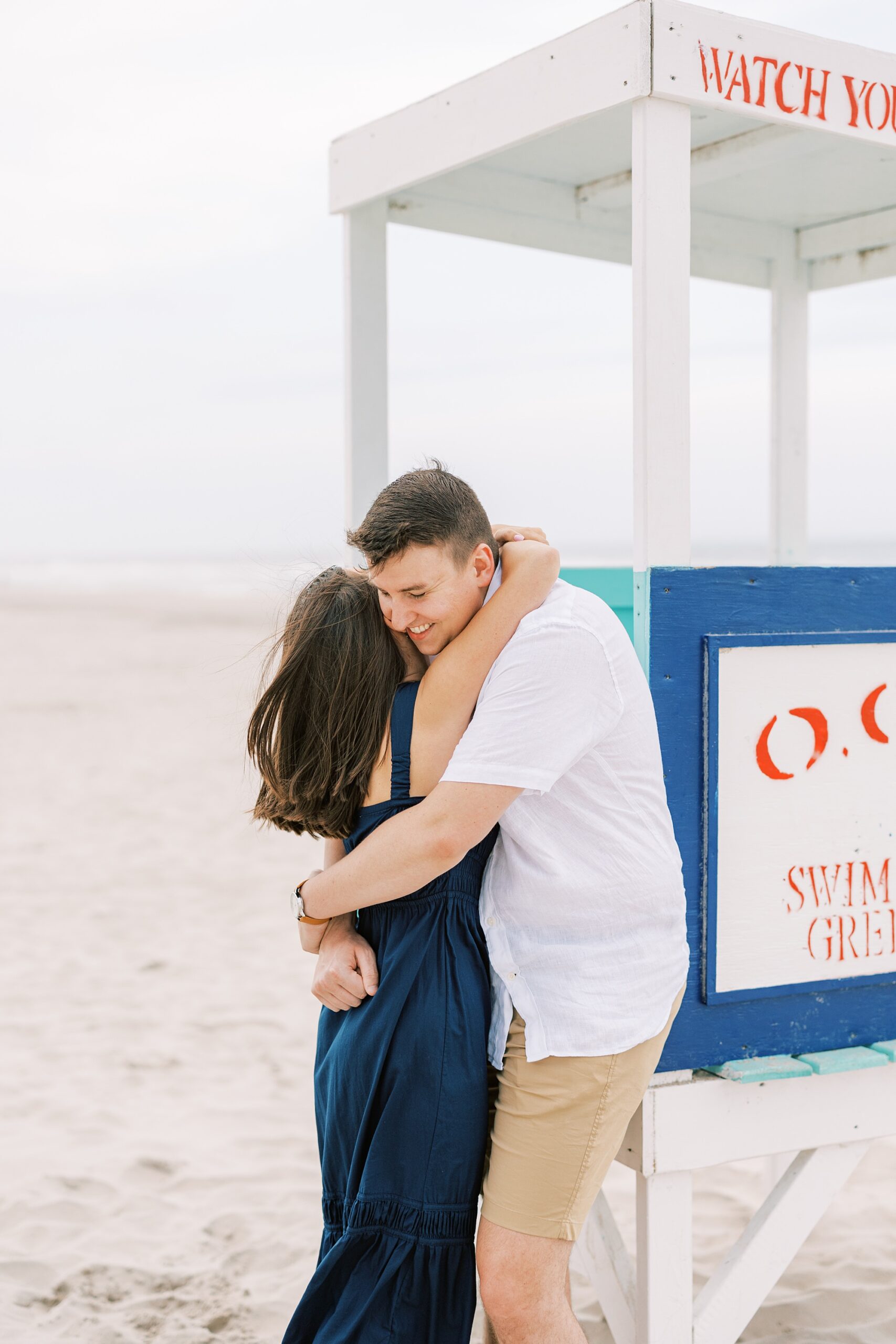 couple hugs near lifeguard stand with blue and white sign on NJ beach