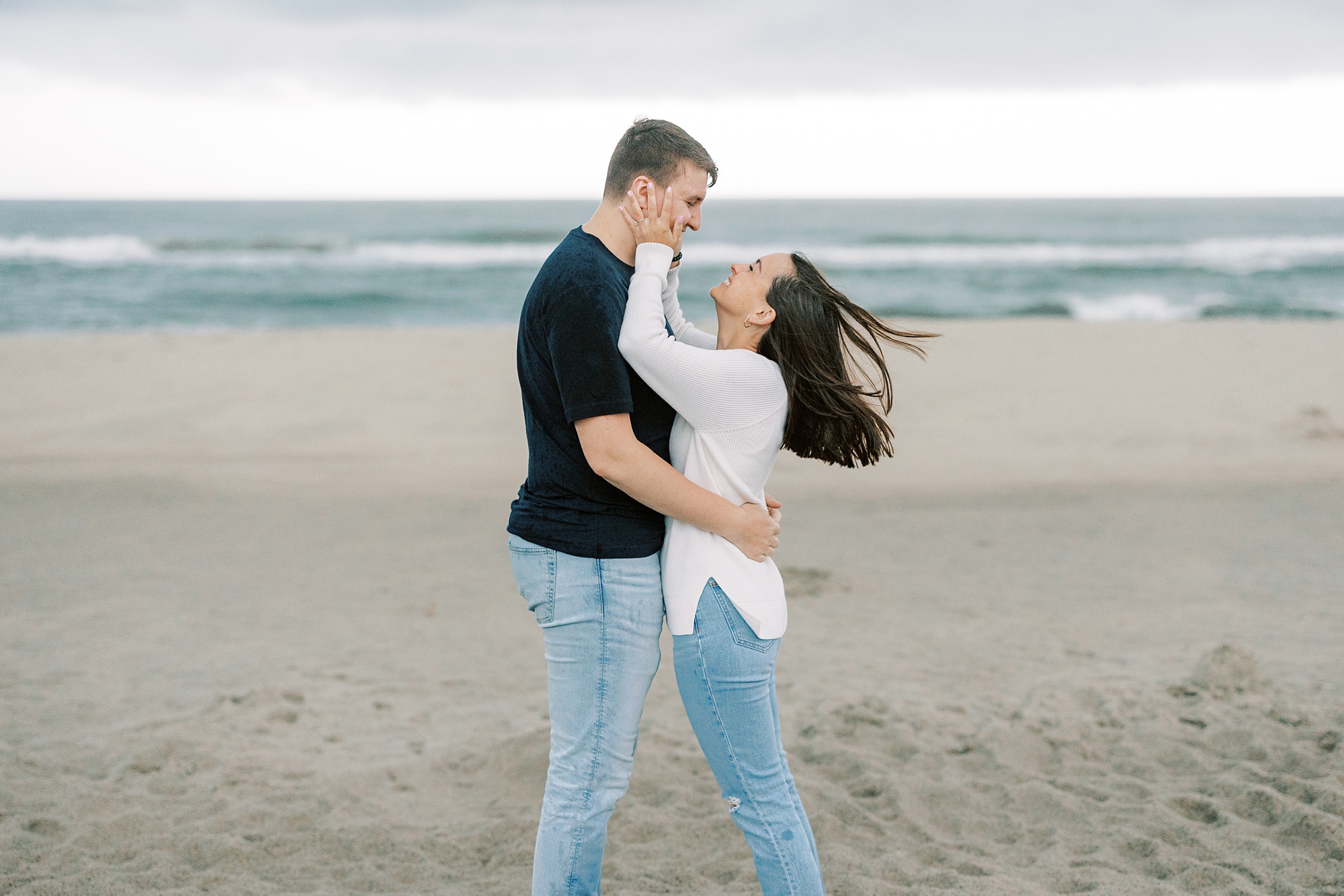 woman in white sweater looks up at man while wind blows their hair on beach 