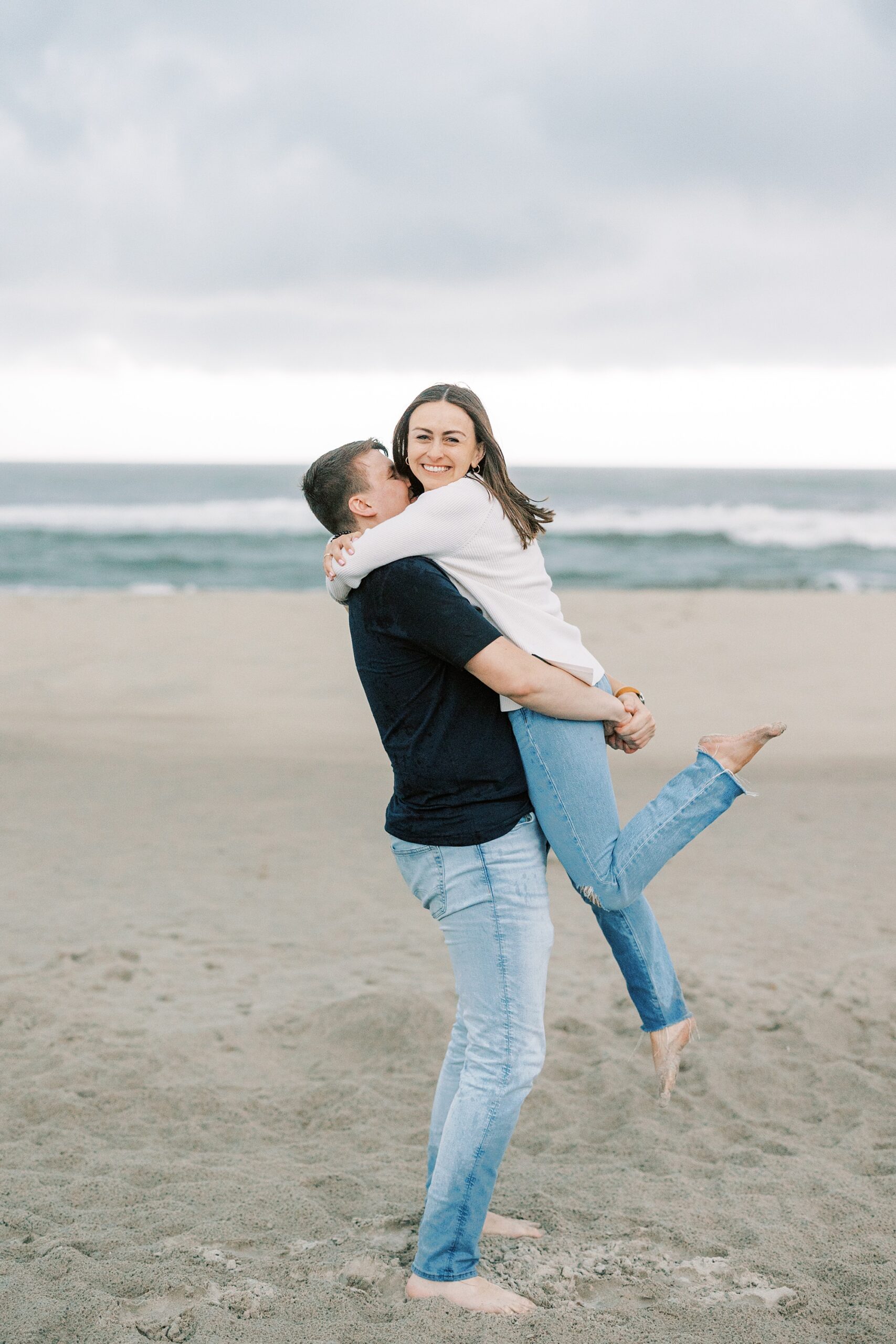 man in black shirt lifts up woman in white sweater on beach during Ocean City NJ engagement photos