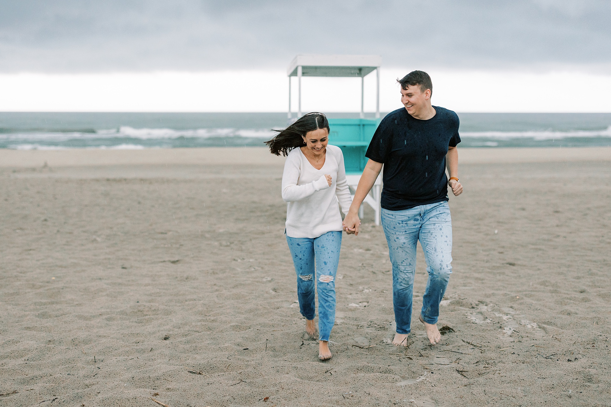 engaged couple holds hands running on beach during rainy Ocean City NJ engagement photos
