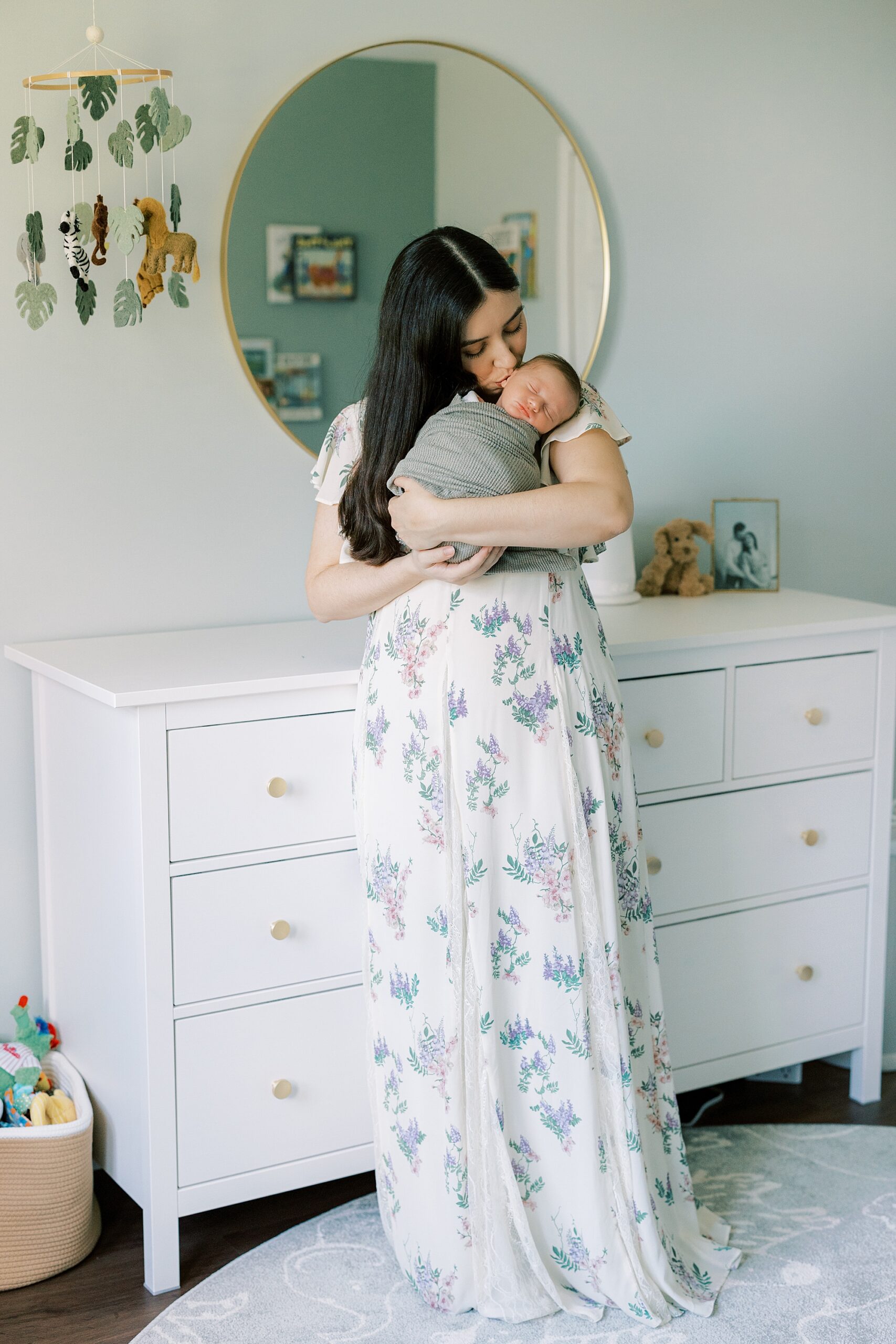 mother in floral gown stands in front of white dresser kissing baby boy on cheek