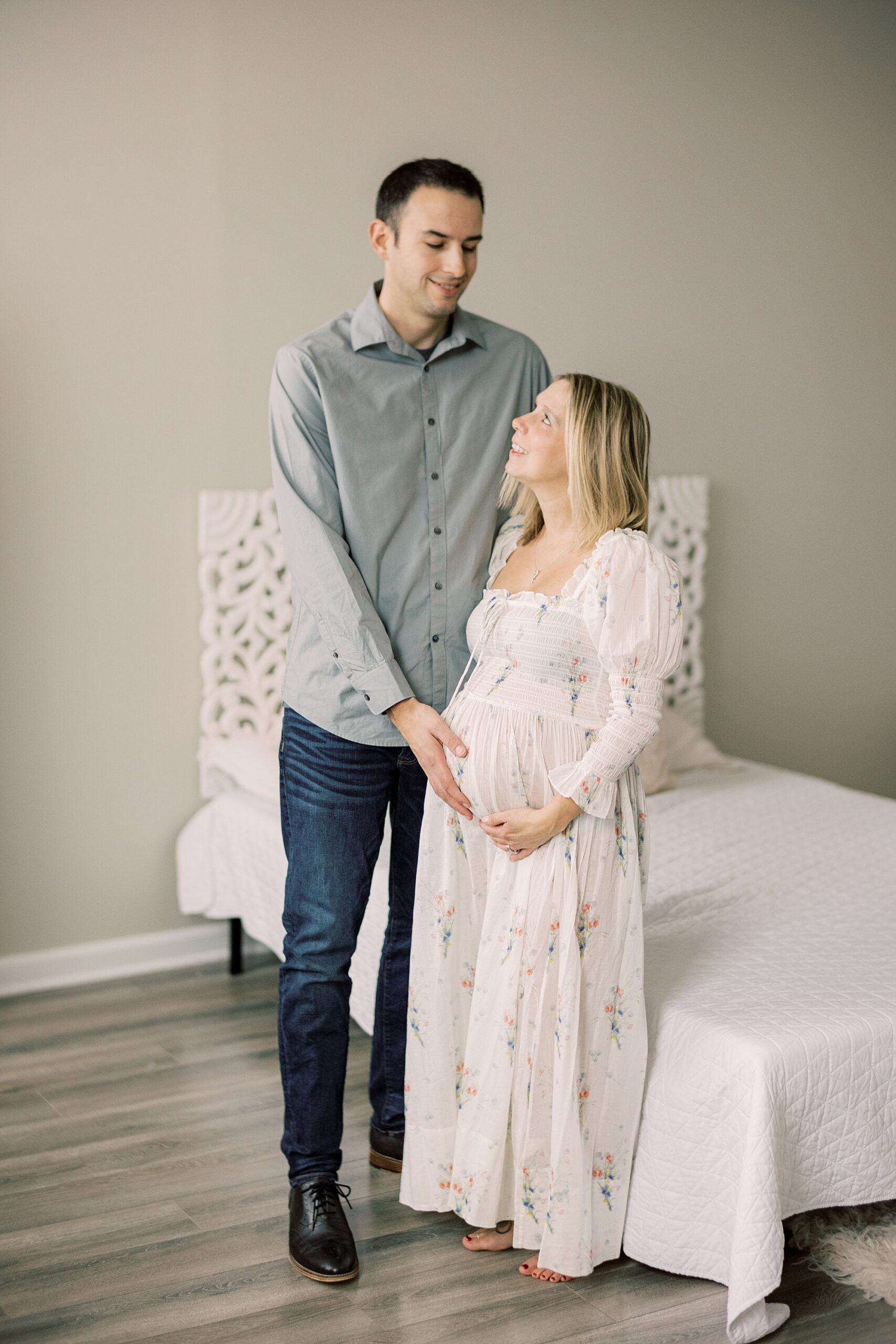 man stands holding wife's baby bump in front of bed during studio maternity session in Media PA