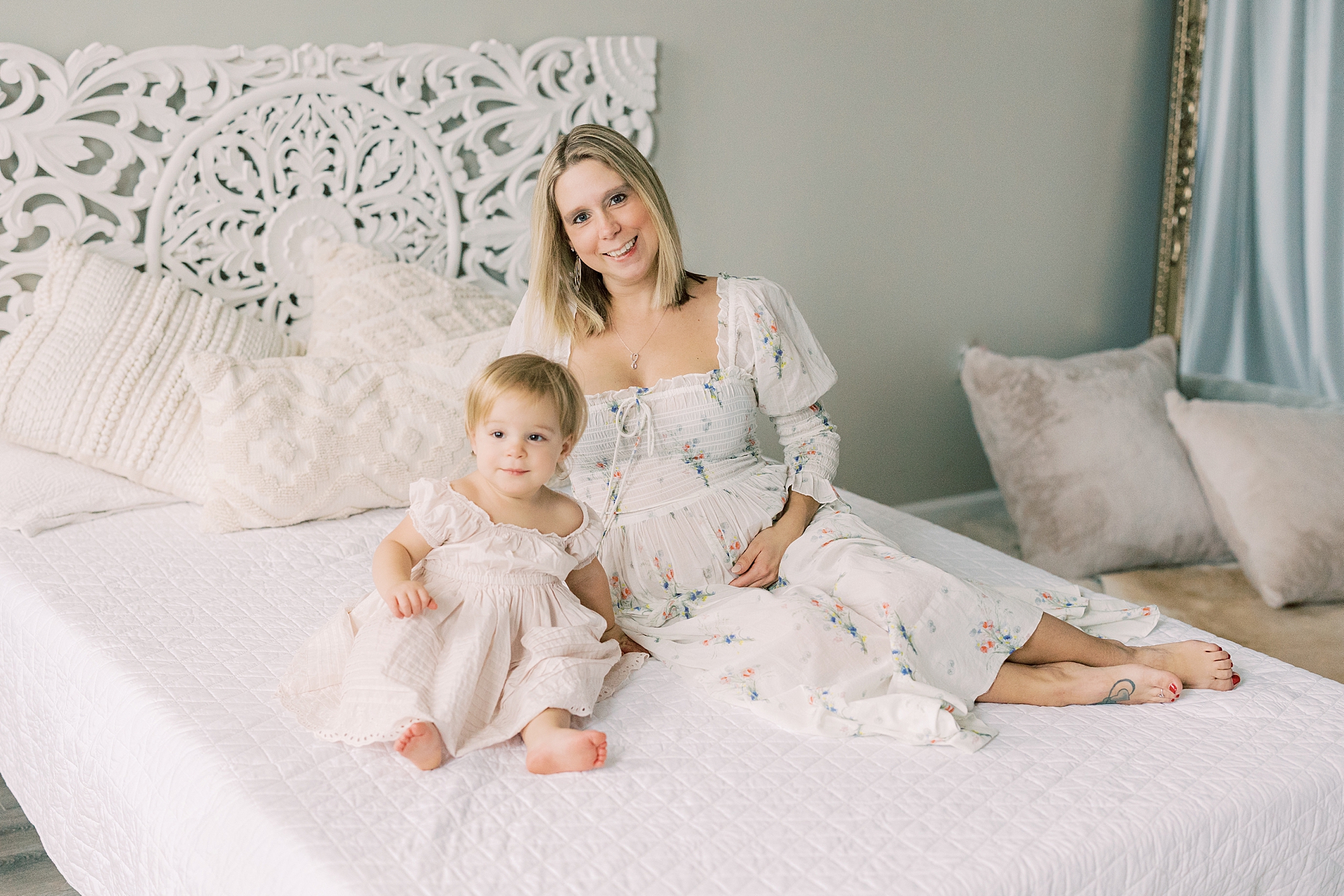 mom sits on white bed holding baby bump and toddler next to her