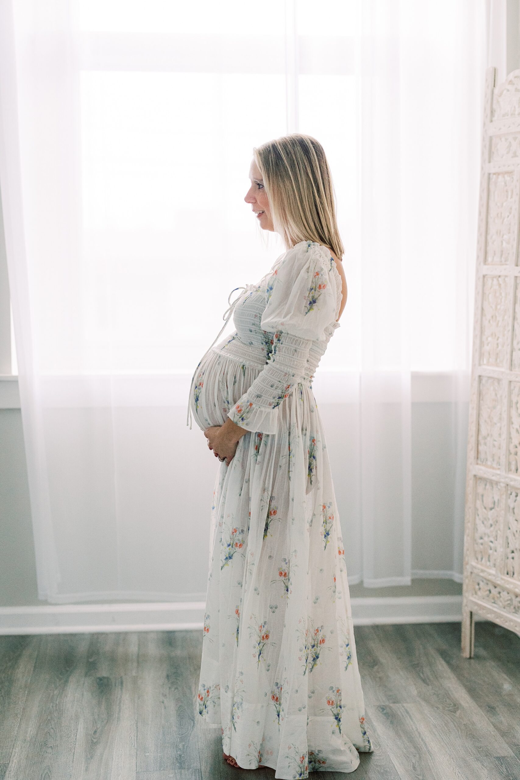 mother stands in window with white curtain holding baby bump
