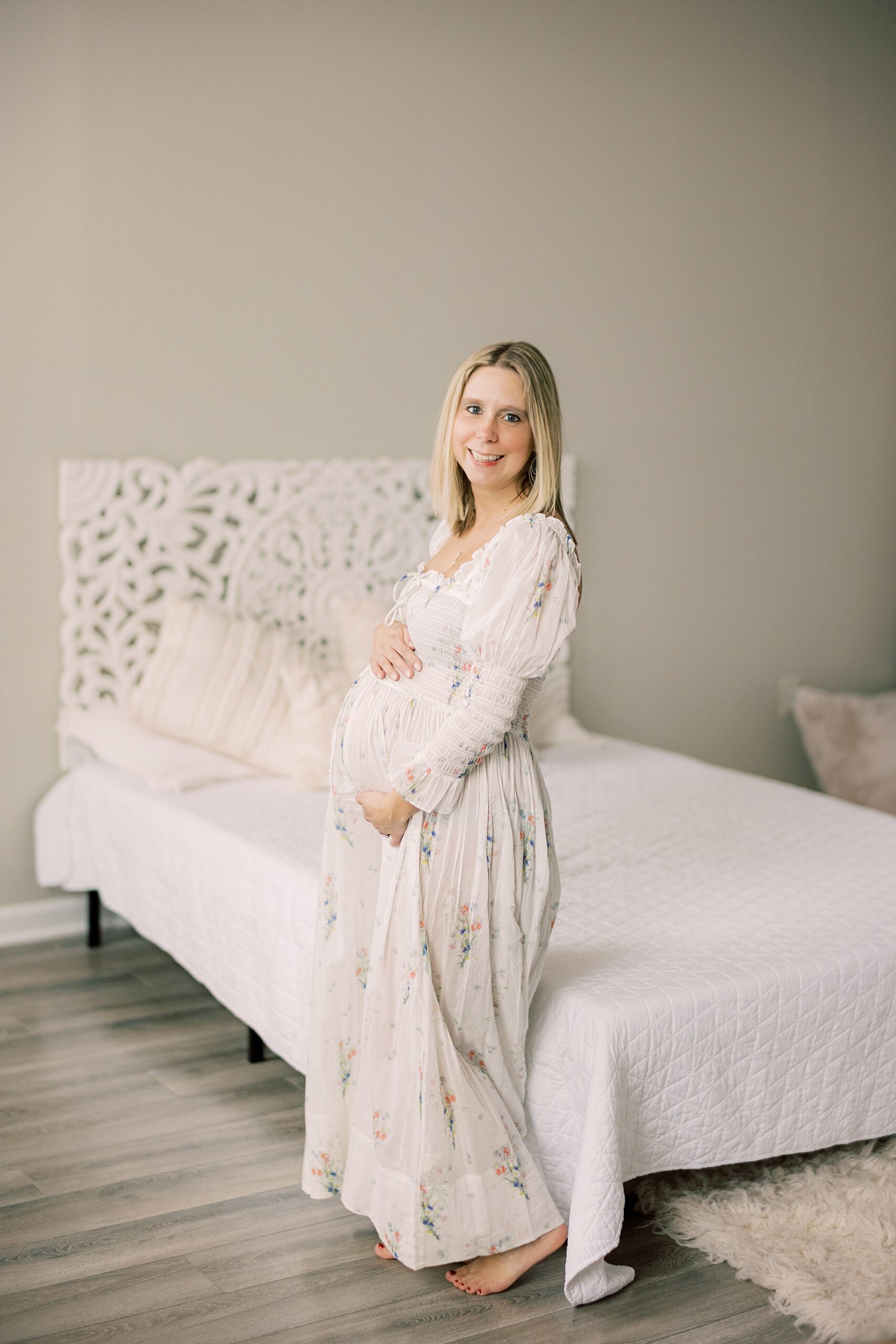 woman stands by white bed holding baby bump during studio maternity session in Media PA