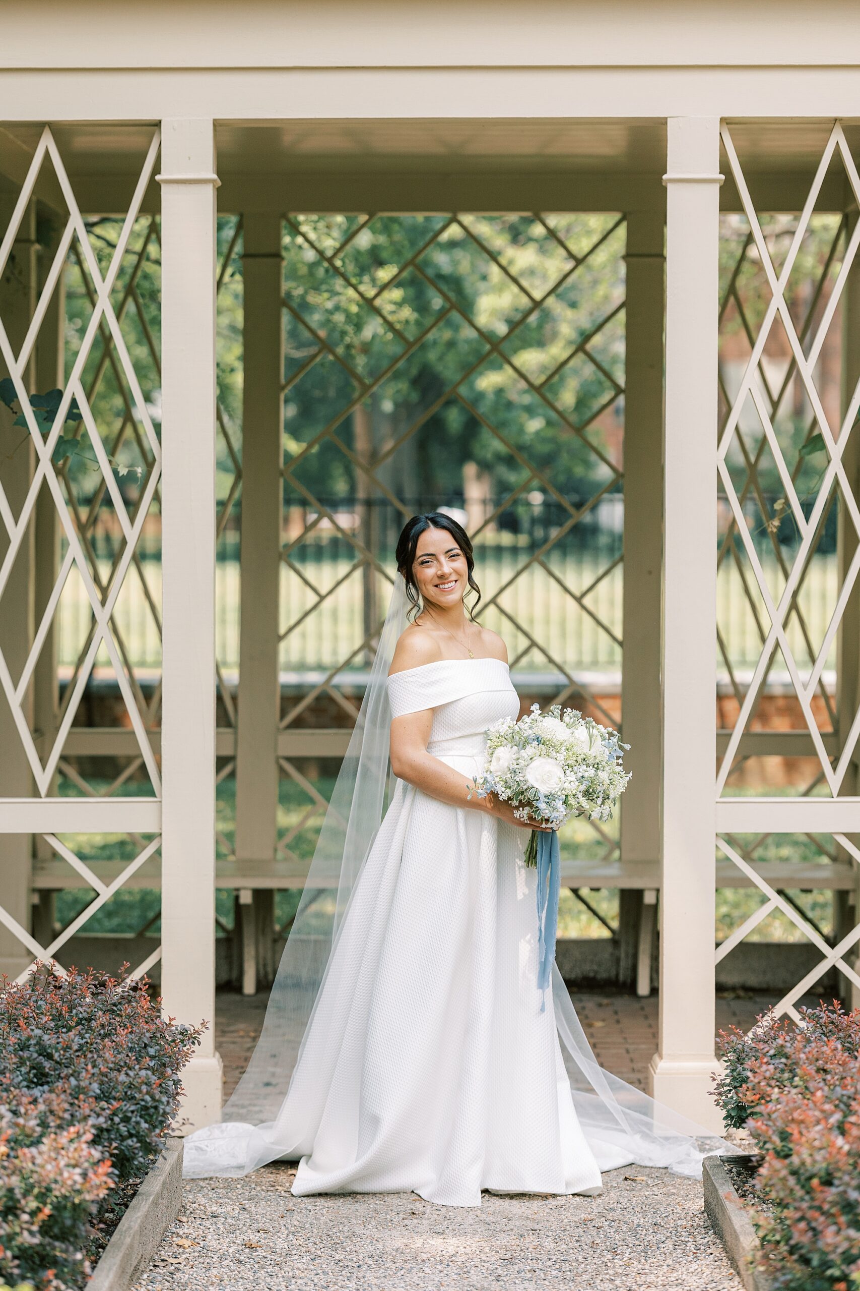 bride in off-the-shoulder gown stands under arbor in Philadelphia PA