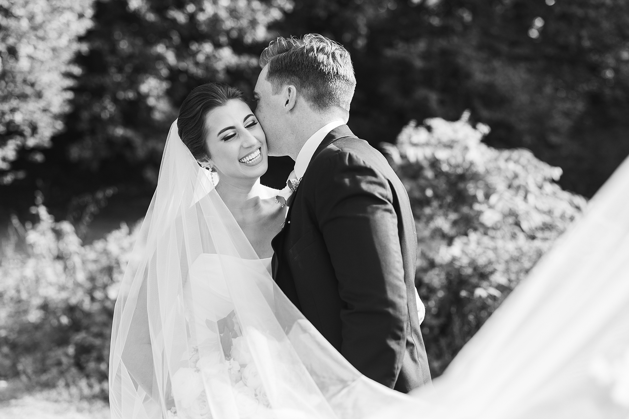 groom leans to kiss bride's cheek with veil floating behind them