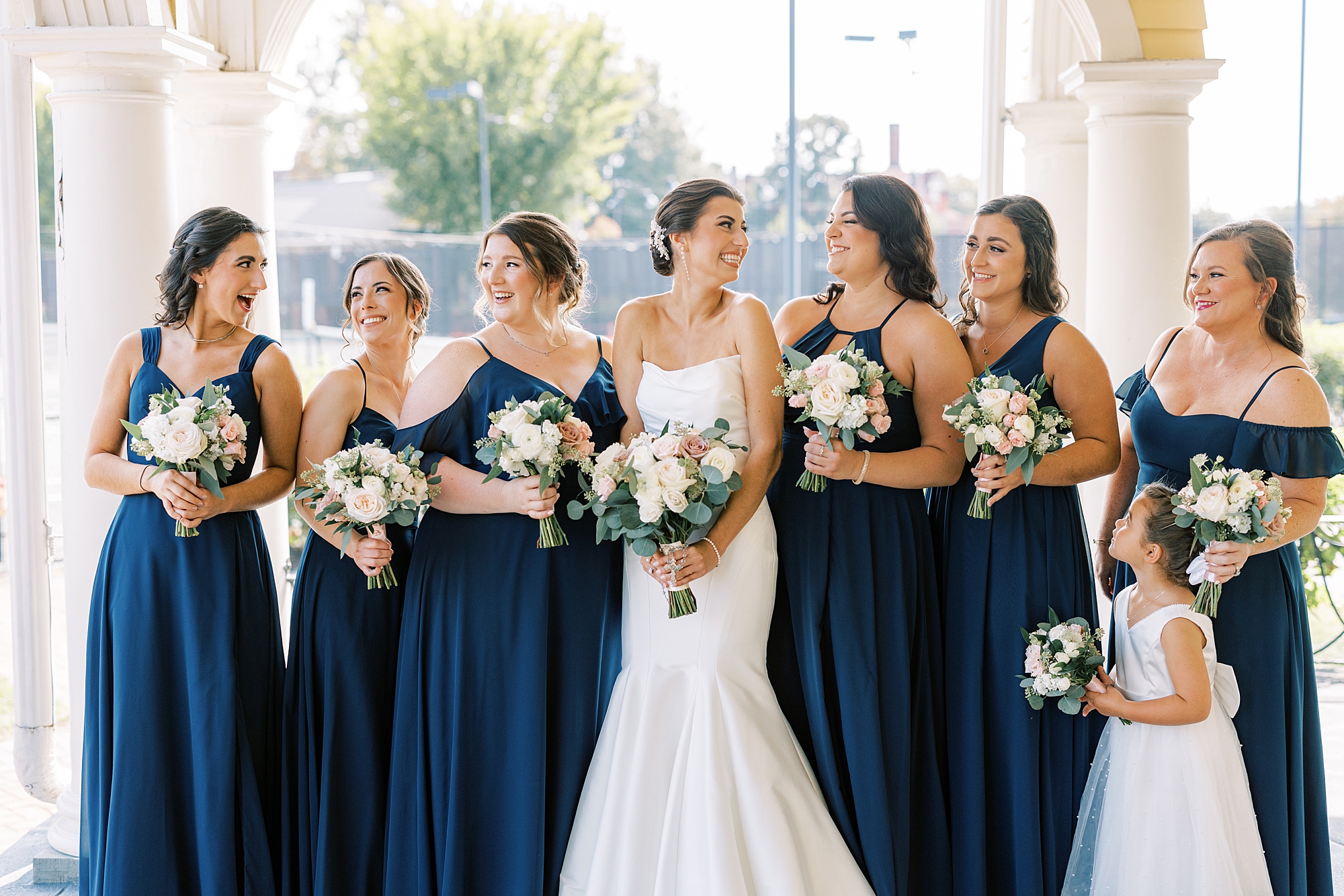 bride smiles with bridesmaids in navy gowns holding bouquet of pink and white flowers 
