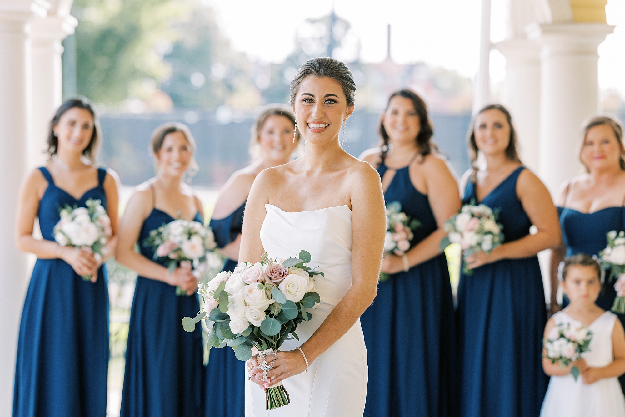 bride smiles holding bouquet of pastel flowers in front of bridesmaids in navy gowns 