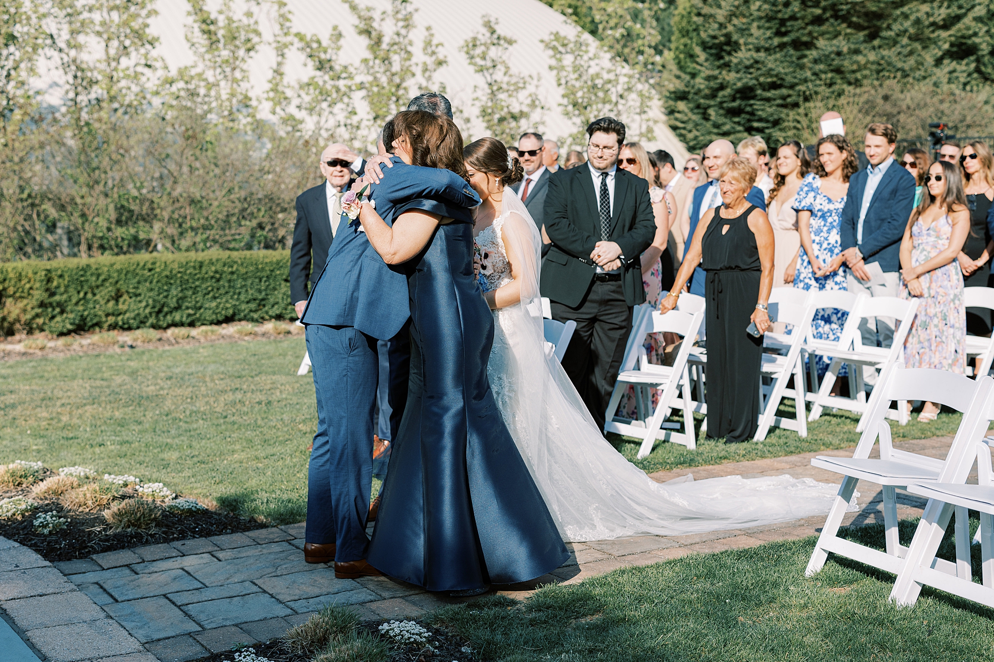 groom hugs mother and father of bride during ceremony in the gardens at Lake House Inn with pink and purple flower installation