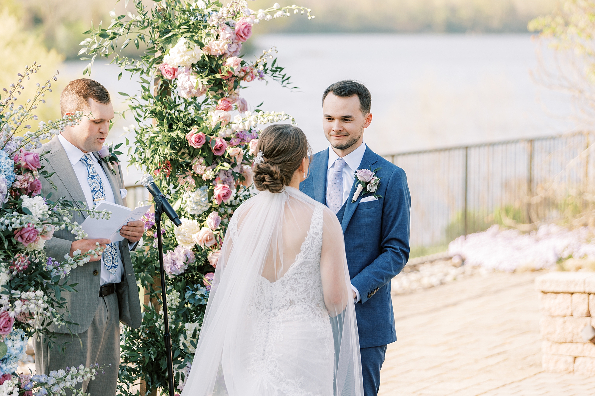 groom looks at bride during ceremony in the gardens at Lake House Inn with pink and purple flower installation