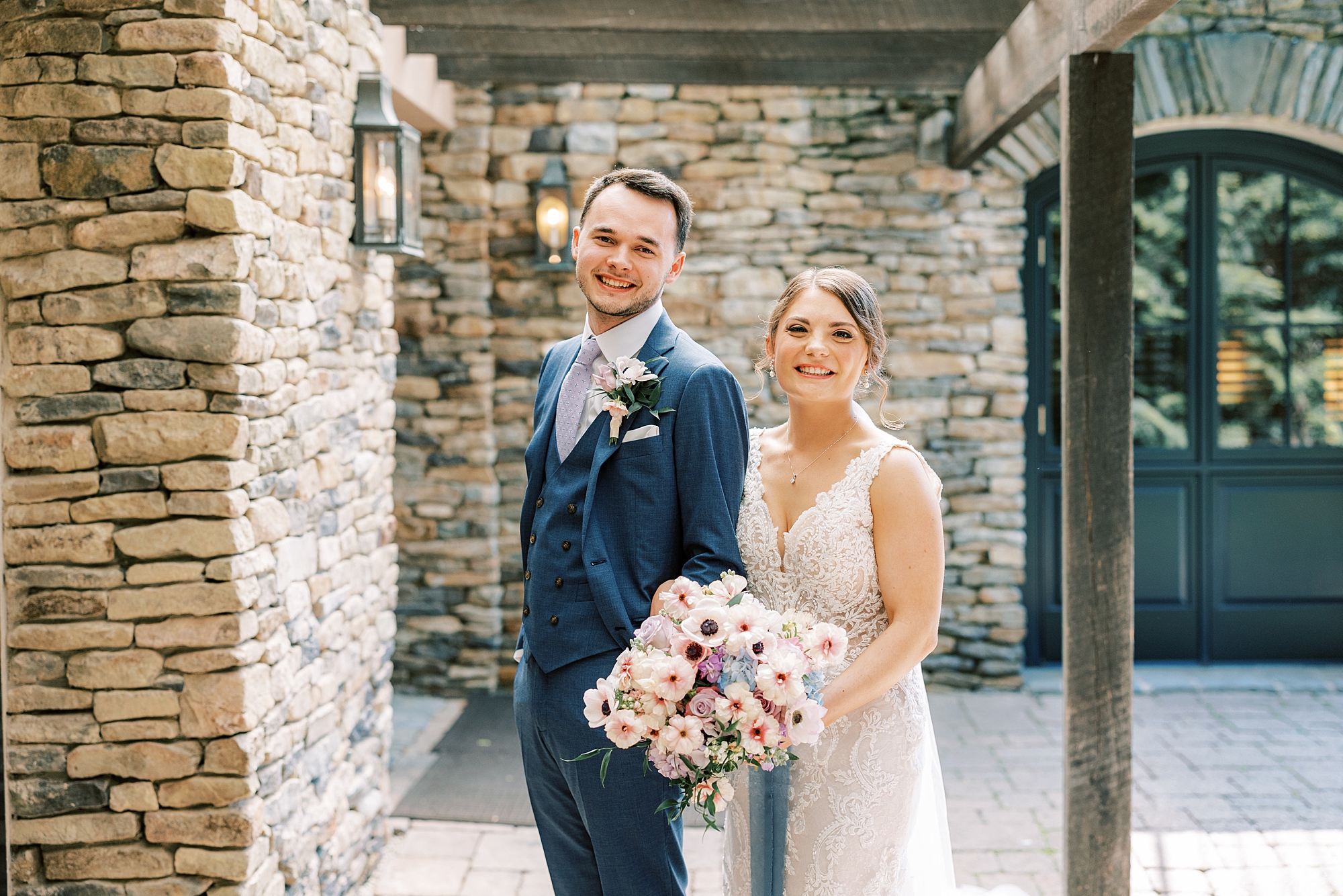 bride stands behind groom smiling during portraits near stone wall at Lake House Inn