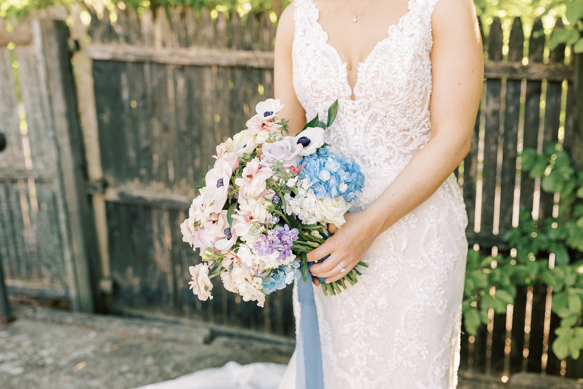 bride holds bouquet of white, pink, and blue flowers near wooden fence 