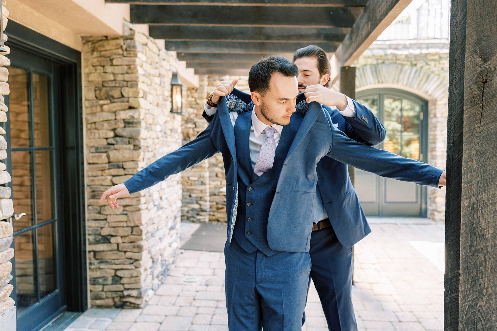 groomsman helps groom into blue suit outside Lake House Inn