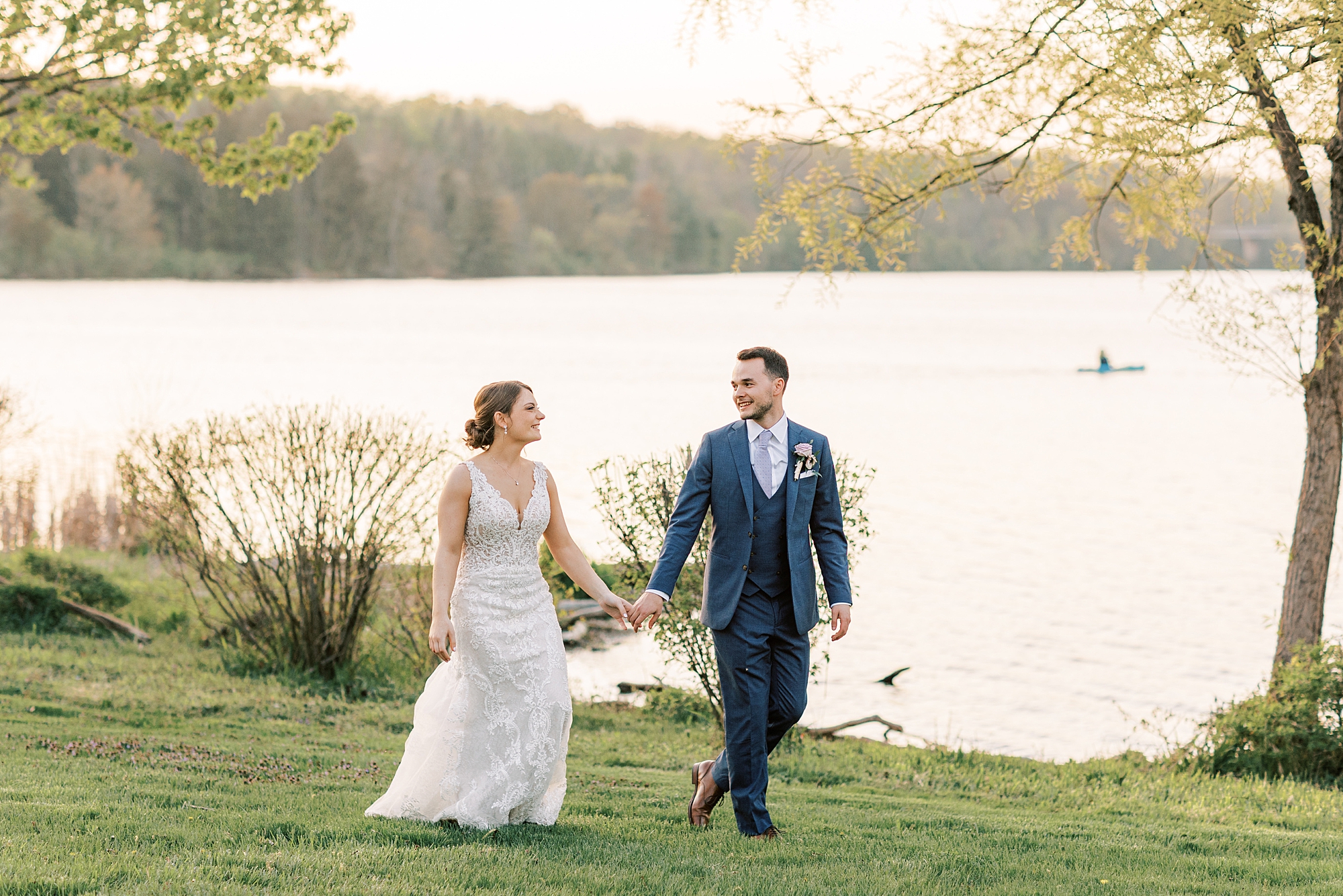 bride and groom hold hands walking near lake at sunset 