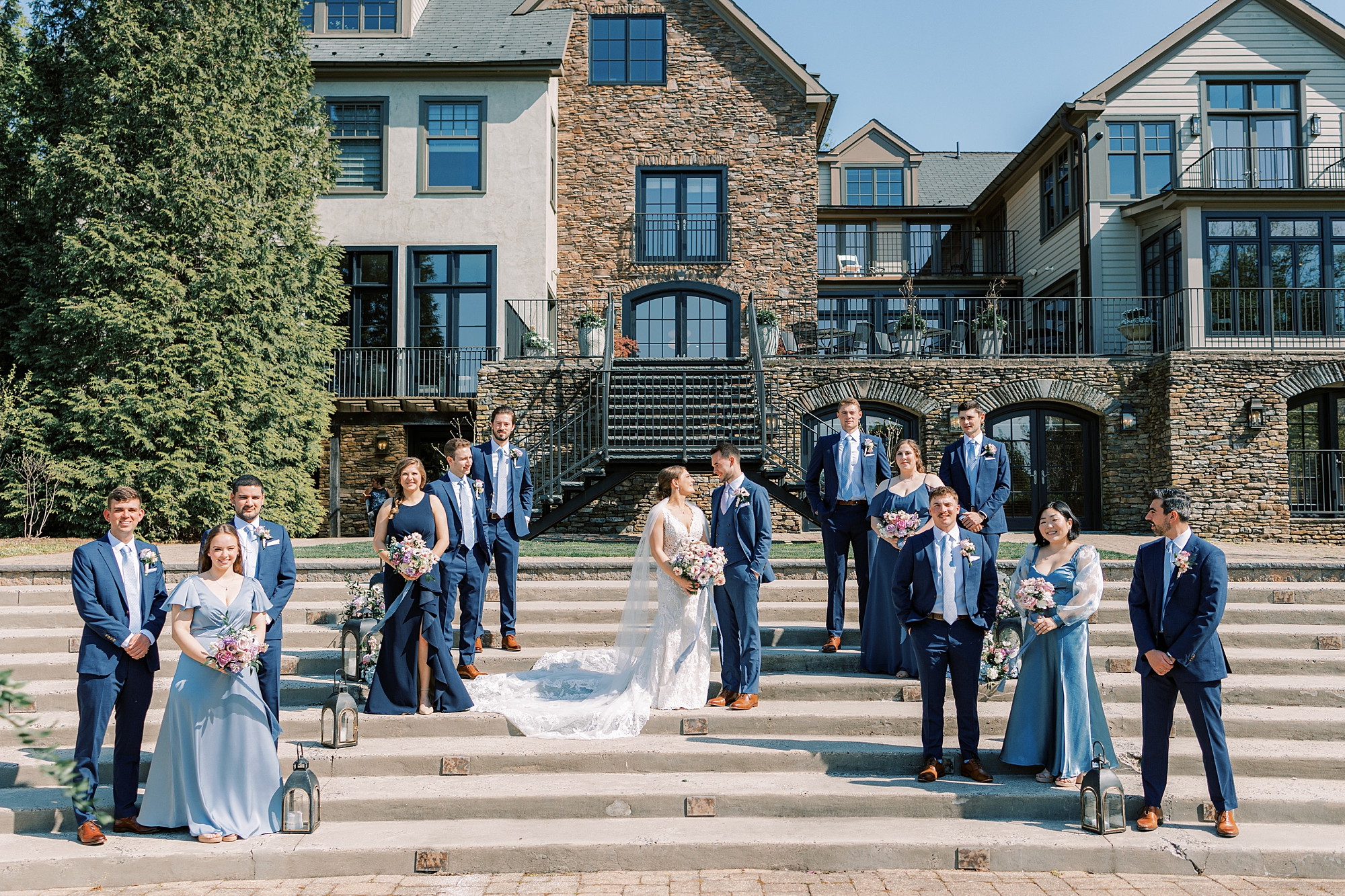 bride and groom kiss on steps of Lake House Inn with wedding party in blue dresses and suits around them