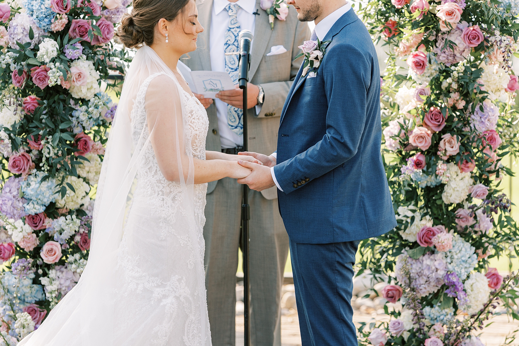 couple holds hands during ceremony in the gardens at Lake House Inn with pink and purple flower installation