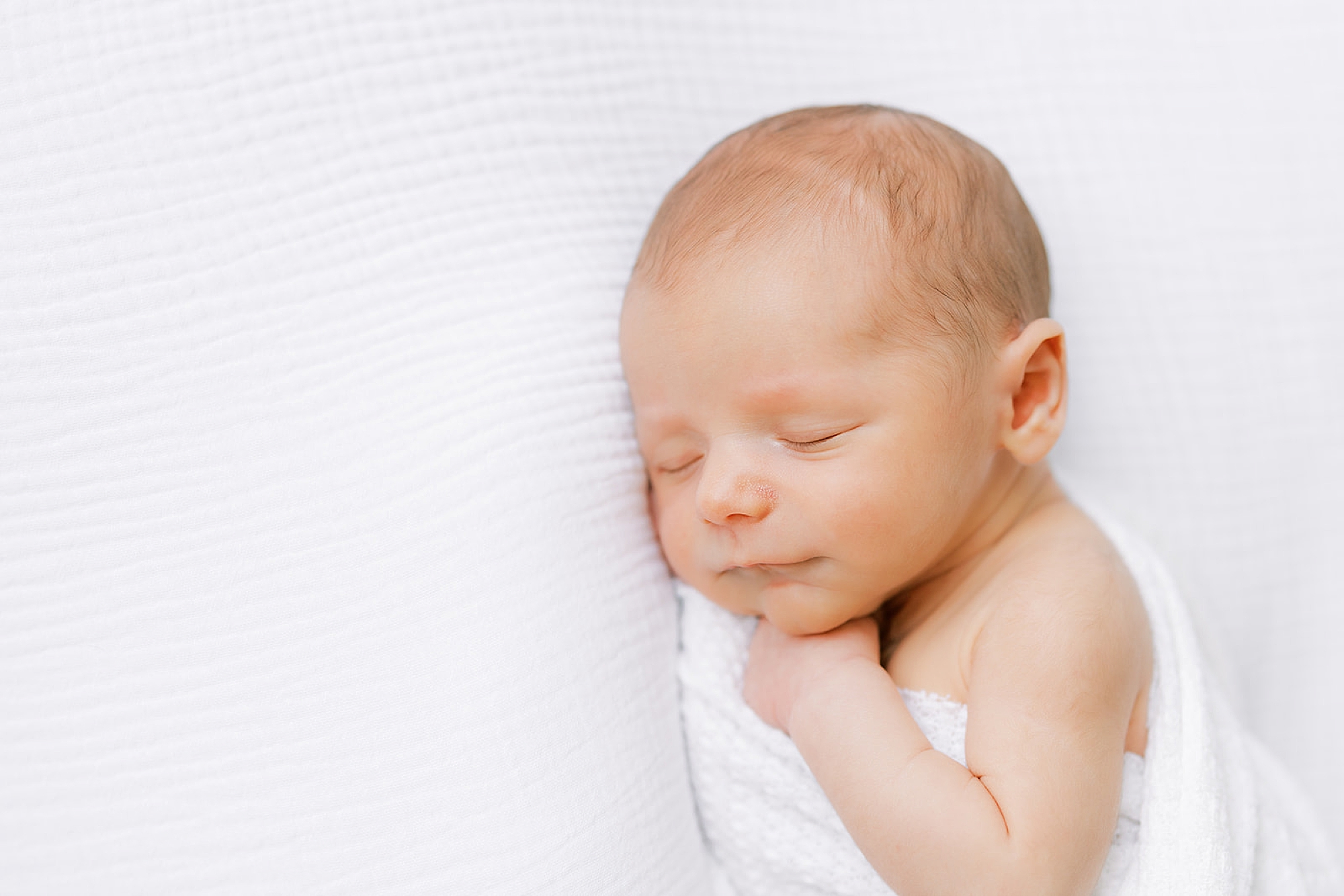 baby lays curled up on white blanket in white wrap during studio newborn photos