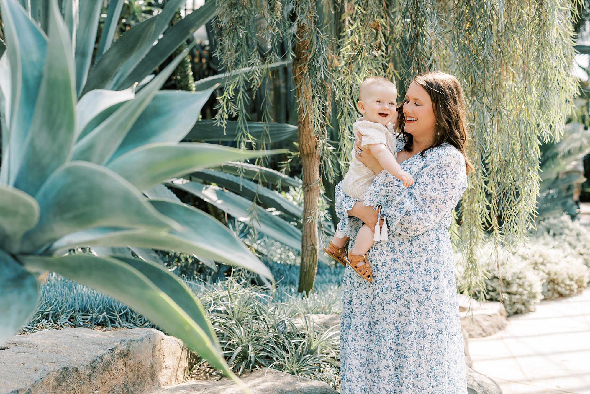 mom smiles at son laughing while she holds him in front of tropical trees 