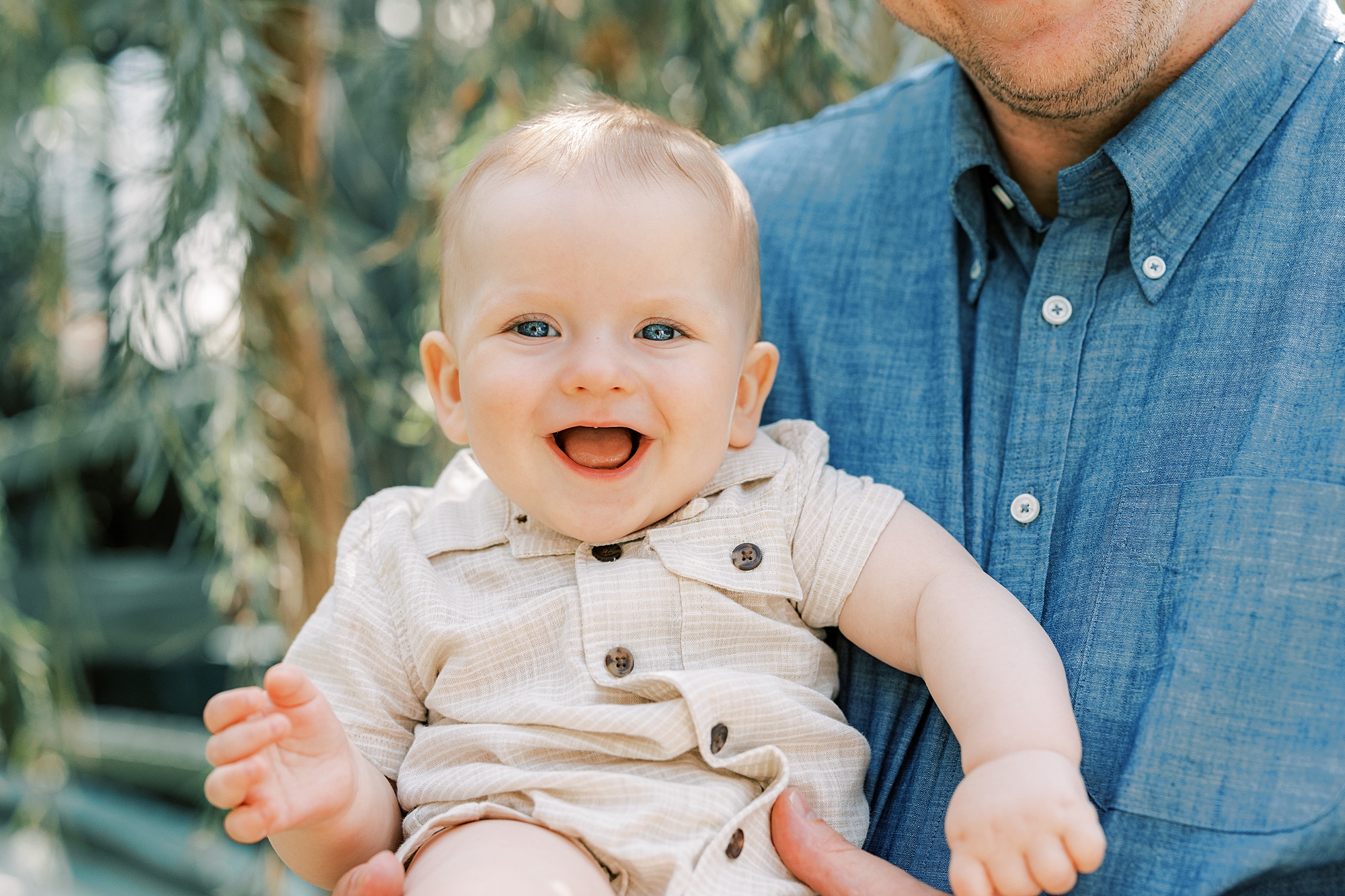 baby boy in tan outfit laughs during family photos at Longwood Gardens