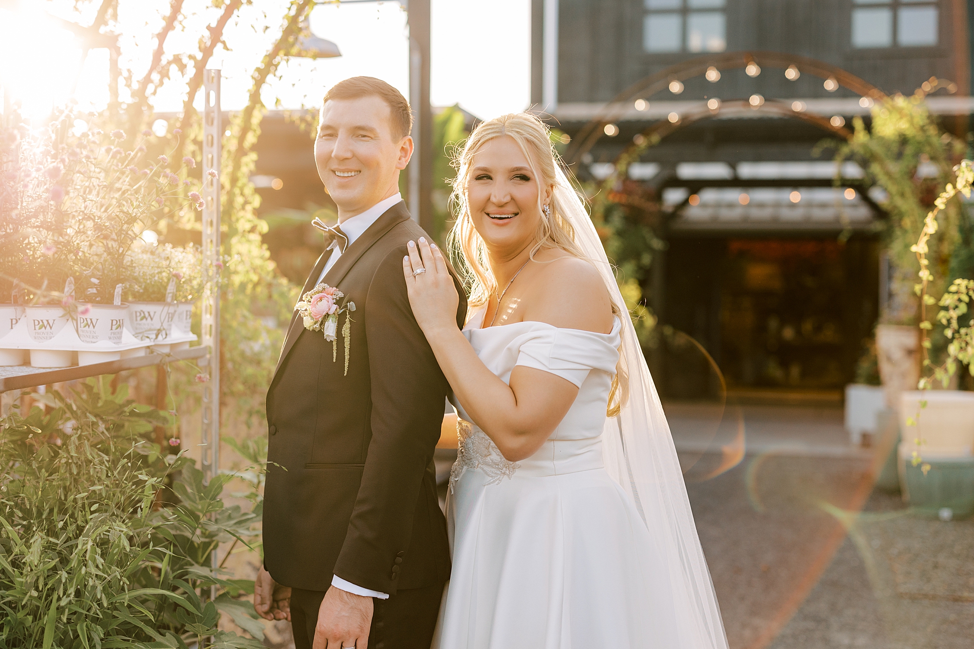 bride stands behind groom holding his shoulder smiling at sunset 
