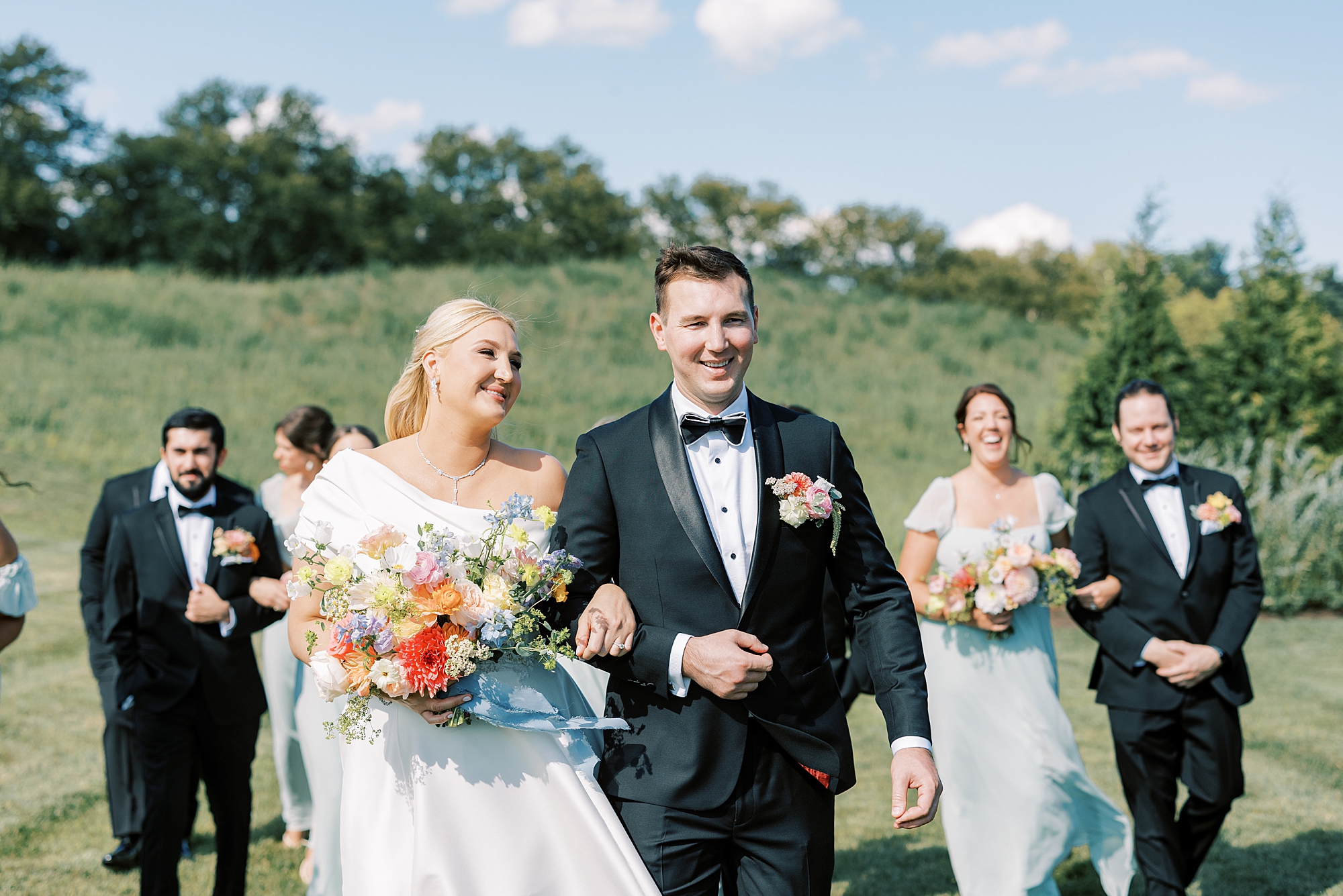 bride smiles at groom walking on field of grass with wedding party behind them 