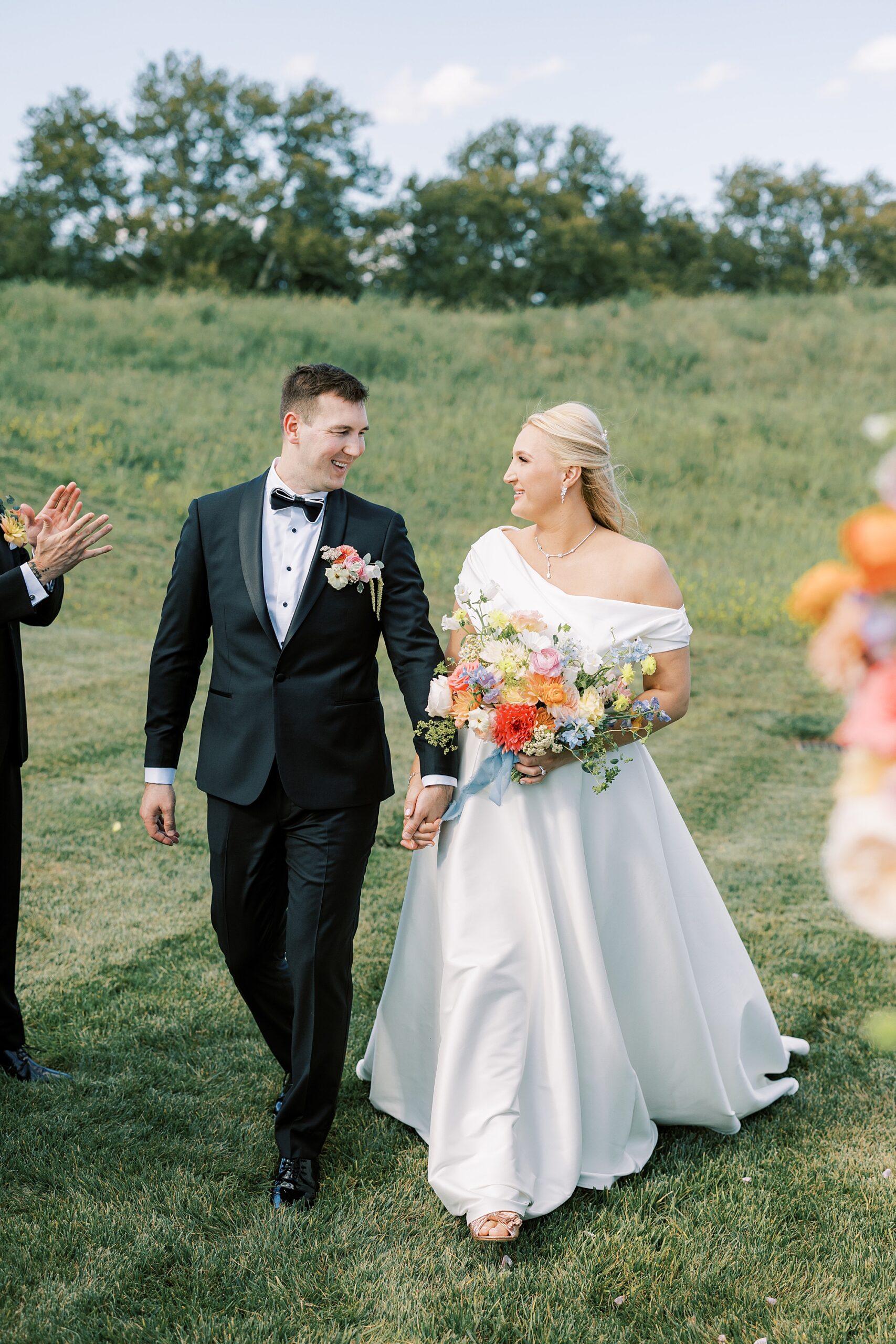 groom in black suit looks over at bride holding her hand walking on lawn at Terrain DelVal