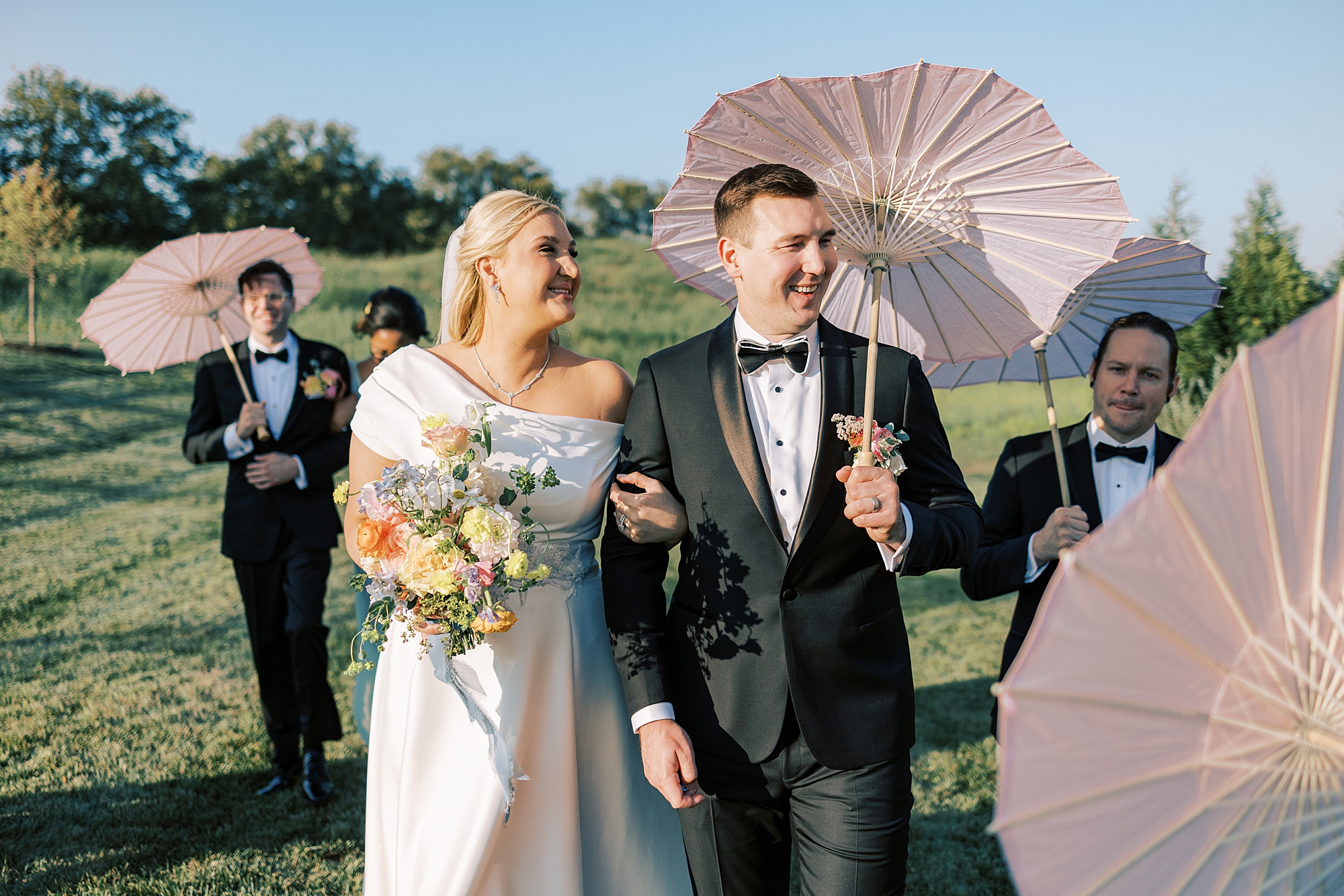 newlyweds laugh walking with wedding party carrying pink parasols 