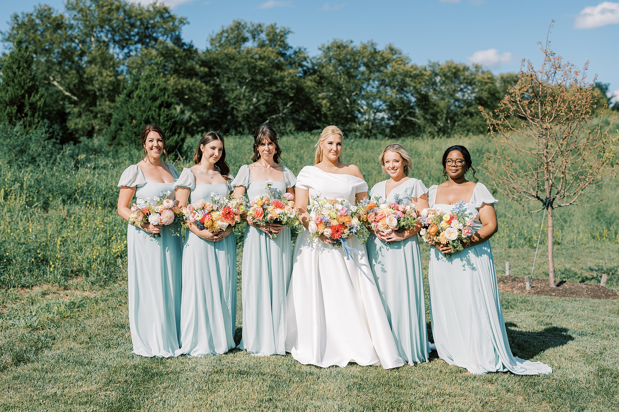 bride poses with bridesmaids in light blue gowns in field at Terrain DelVal