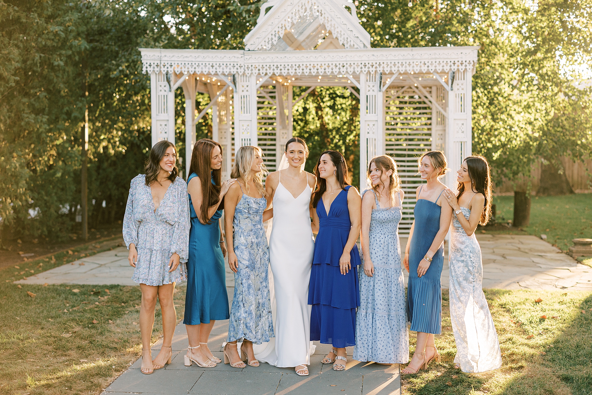 bride stands with bridesmaids in mismatched blue gowns in front of gazebo at the Terrain at Styer's