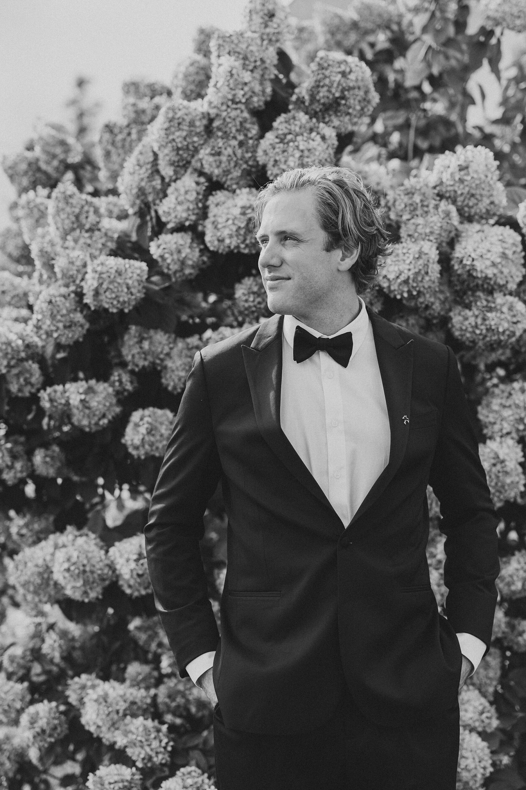 groom stands in front of hydrangea bush at Lauxmont Farms with hand sin pocket 