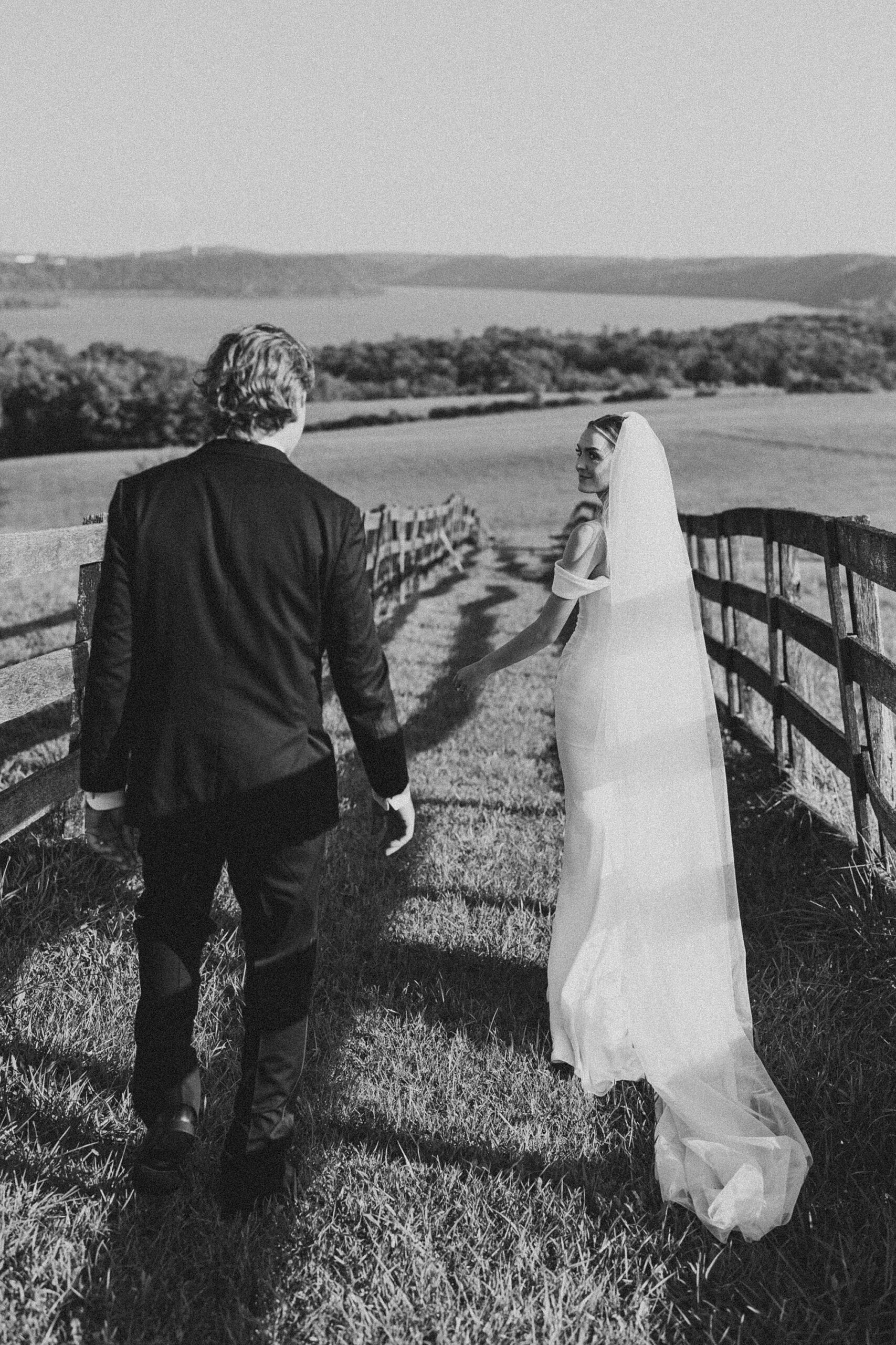 bride and groom hold hands looking back walking between fences