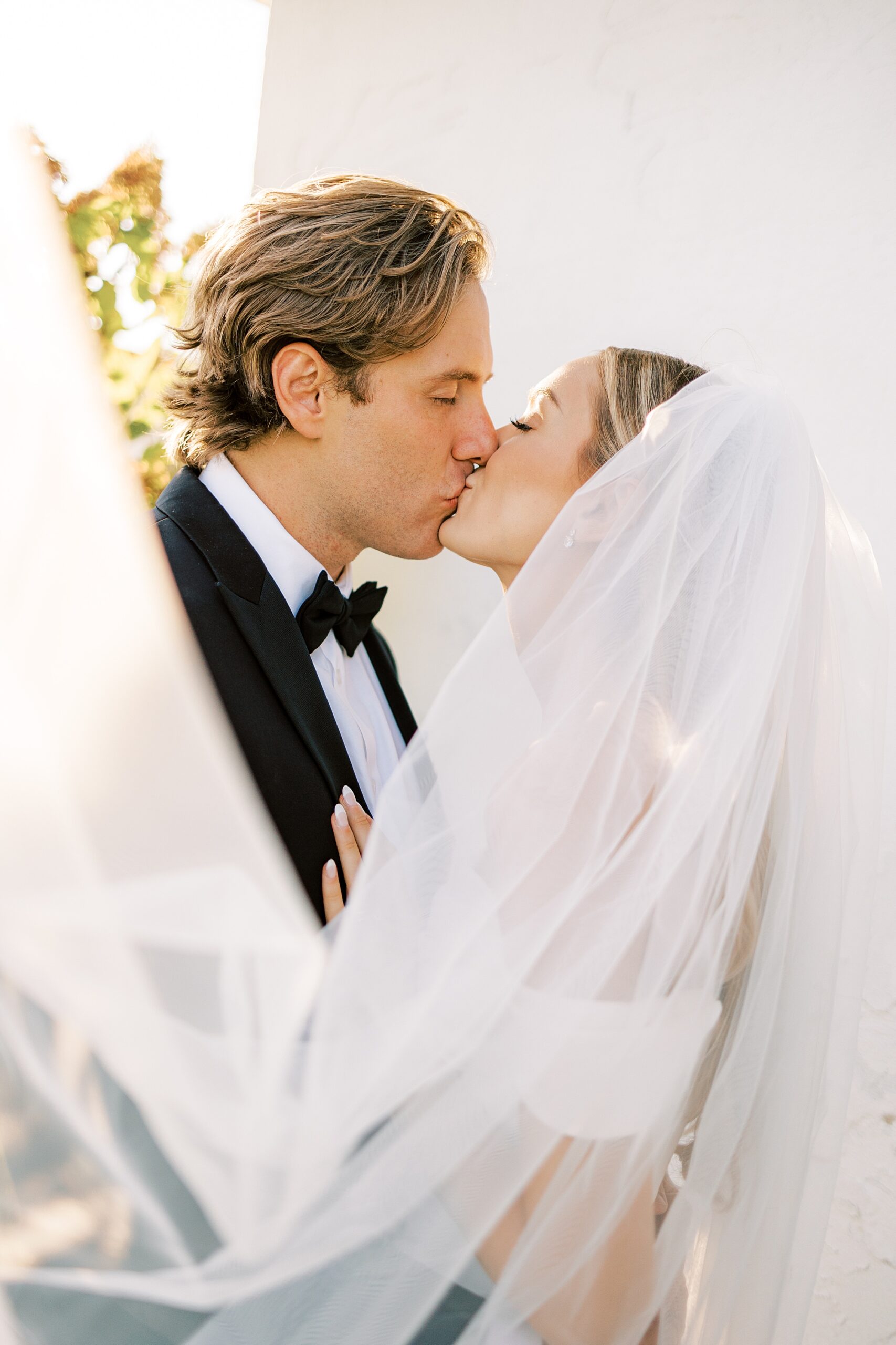 bride and groom kiss with veil lifted up around them outside white wall at Lauxmont Farms