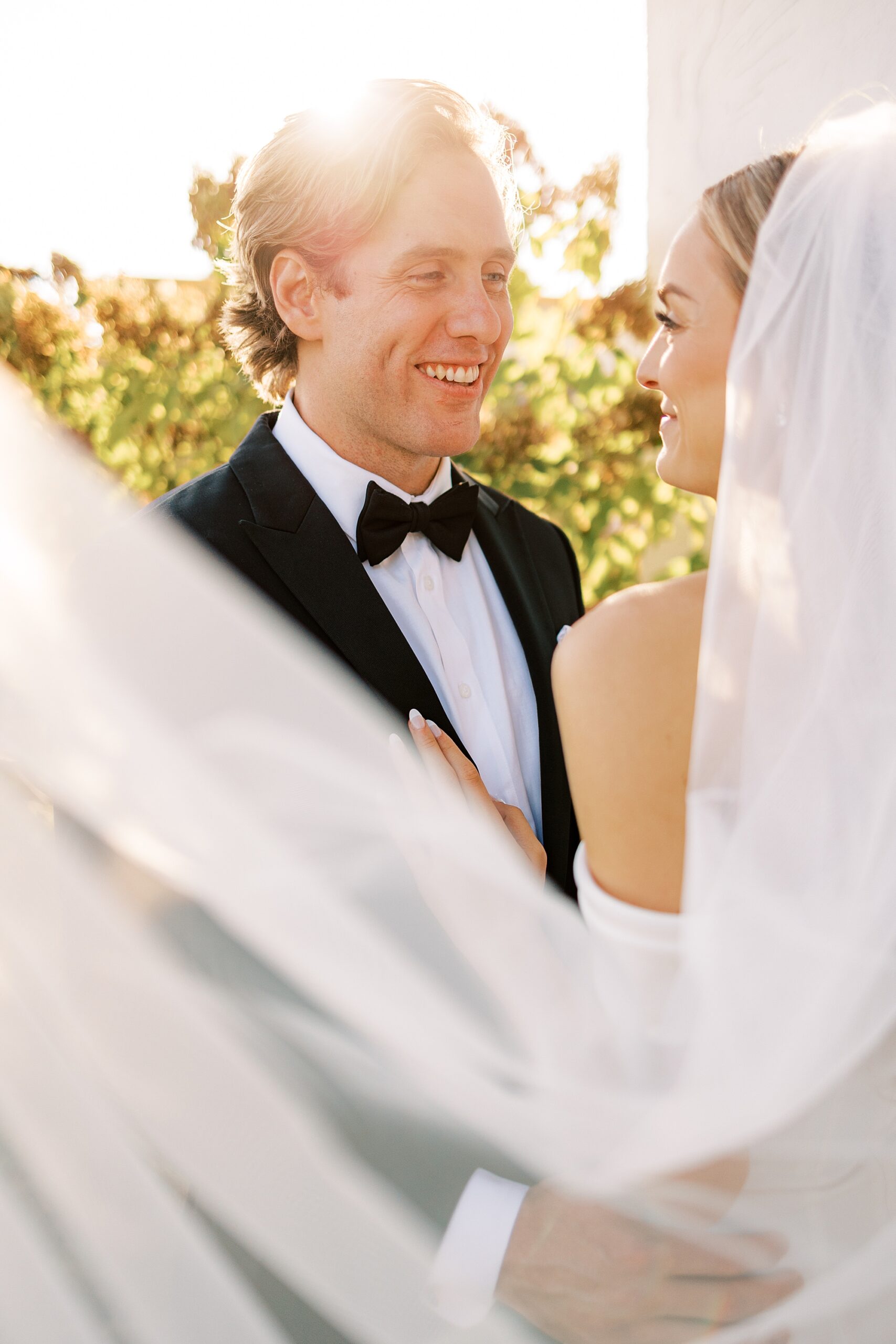 groom in black suit looks at bride outside white wall at Lauxmont Farms