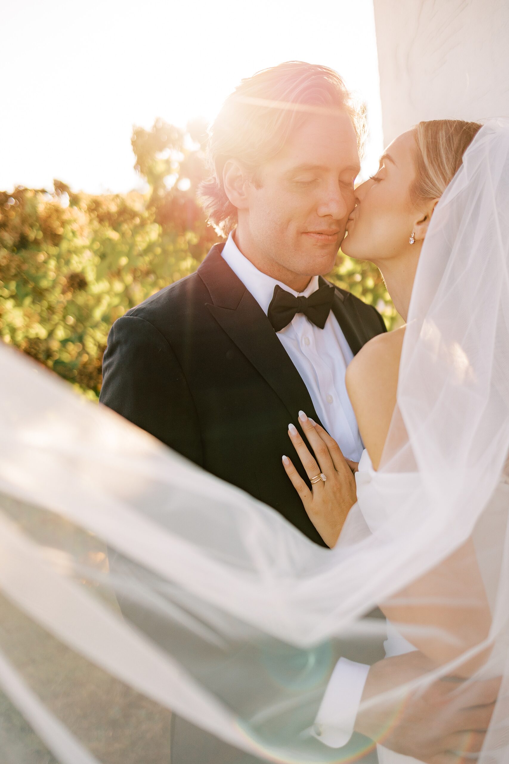 bride leans to kiss groom's cheek on tux jacket with bride's veil floating behind them 