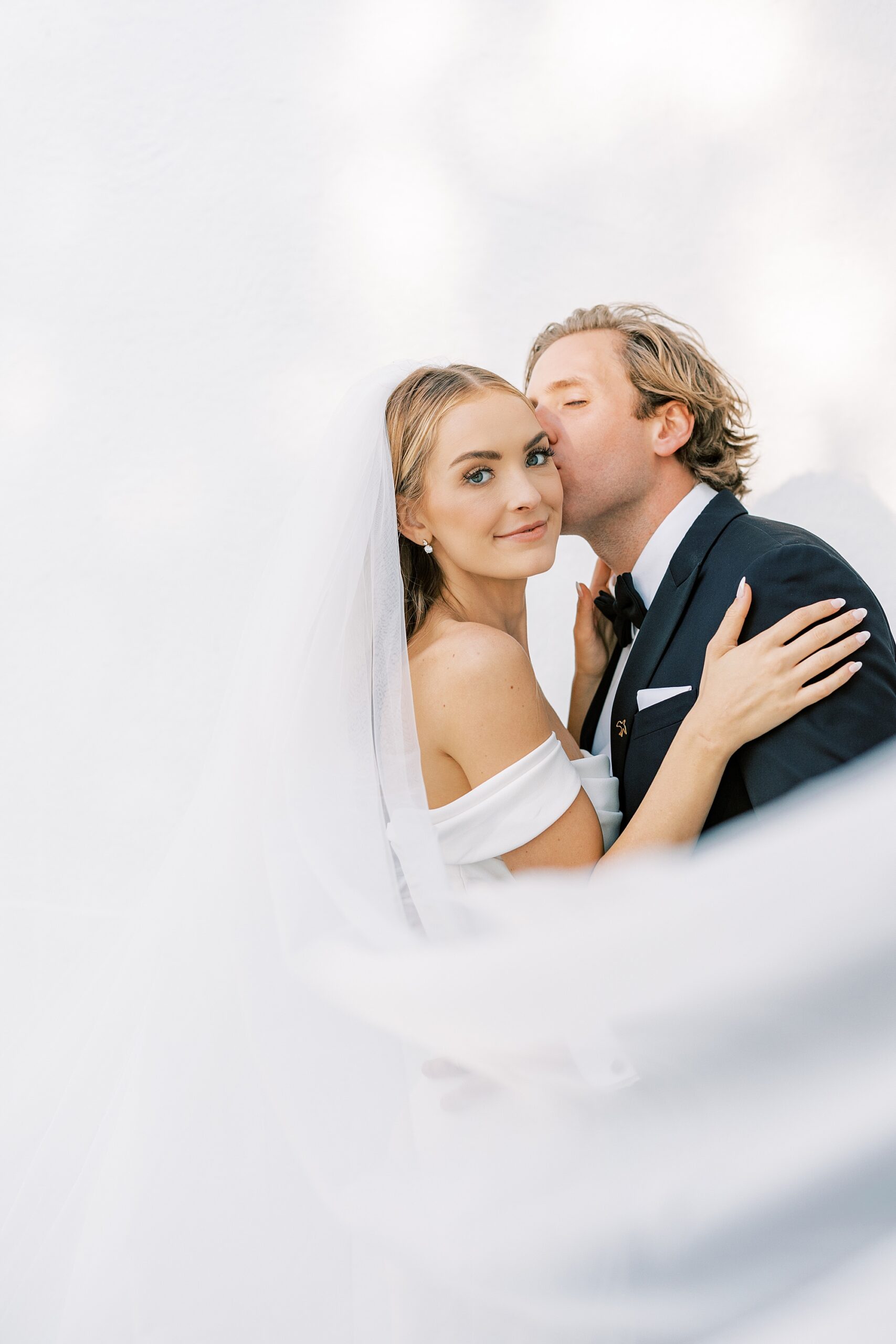 groom kisses bride's cheek with veil floating behind them 