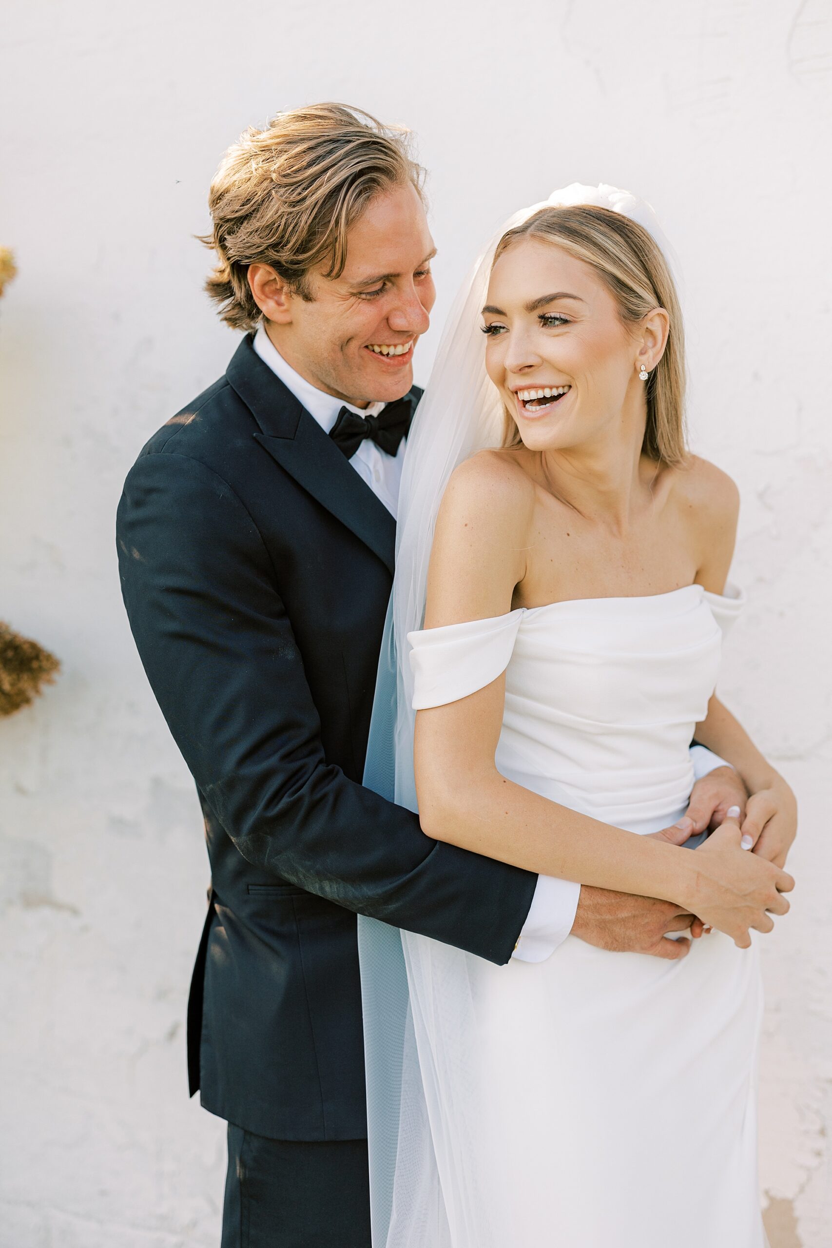 groom hugs bride around middle from behind near white wall at Lauxmont Farms