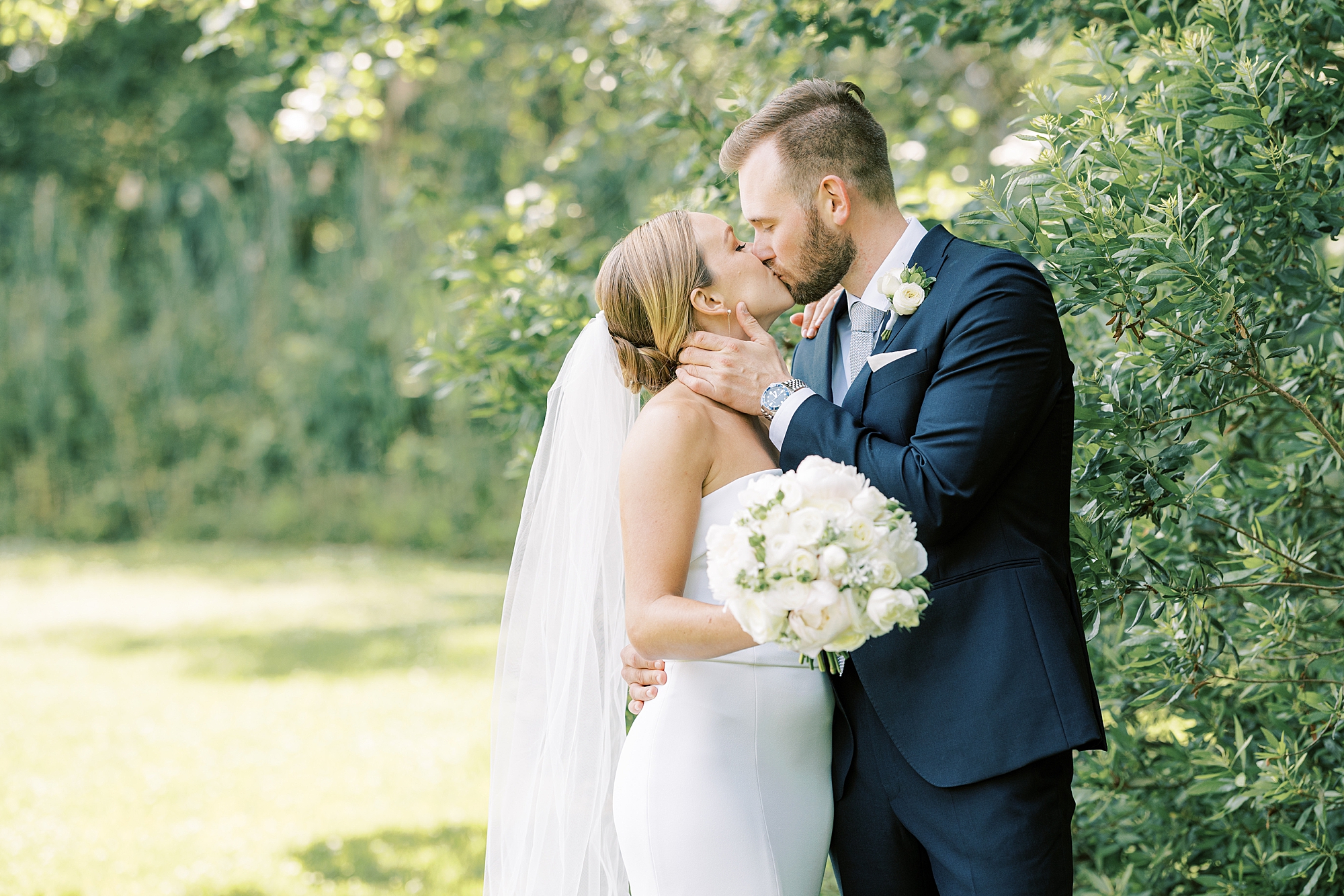 man and woman kiss on lawn at Willow Creek Winery with bouquet of white and ivory flowers 