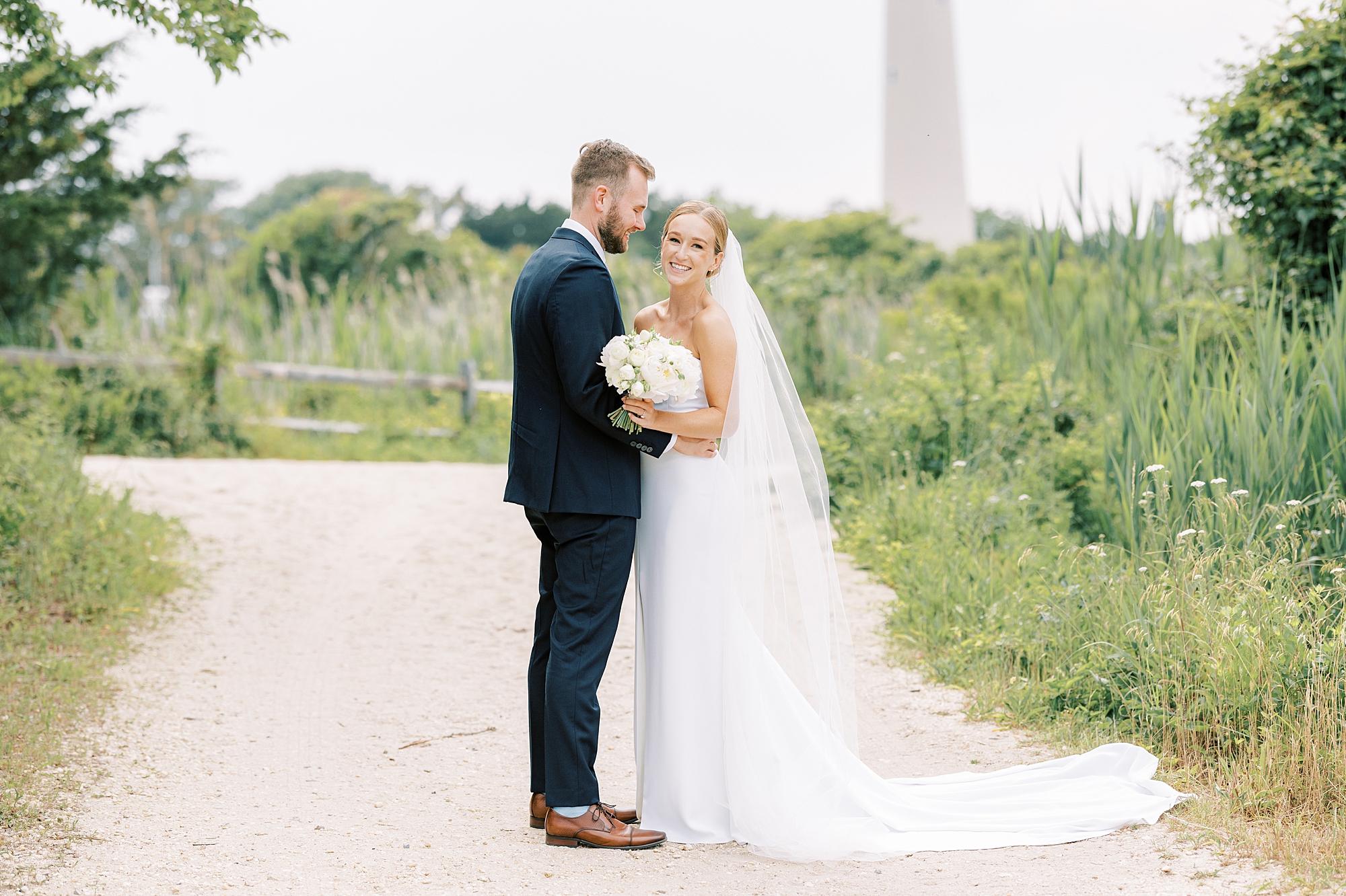 groom looks down at bride hugging her to him in front of the Cape May lighthouse 